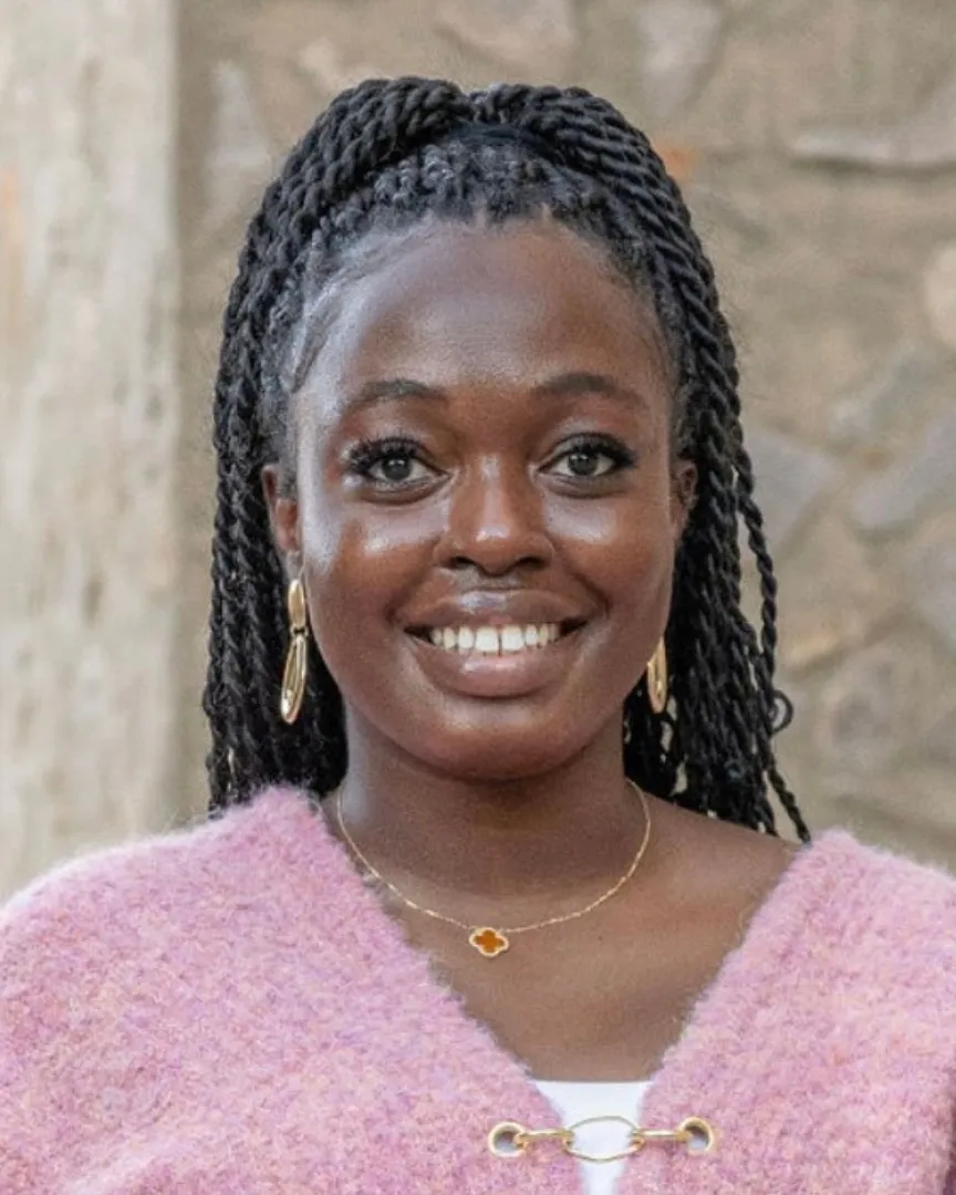 Smiling woman with braided hair wearing a pink sweater and gold jewelry against a stone wall.