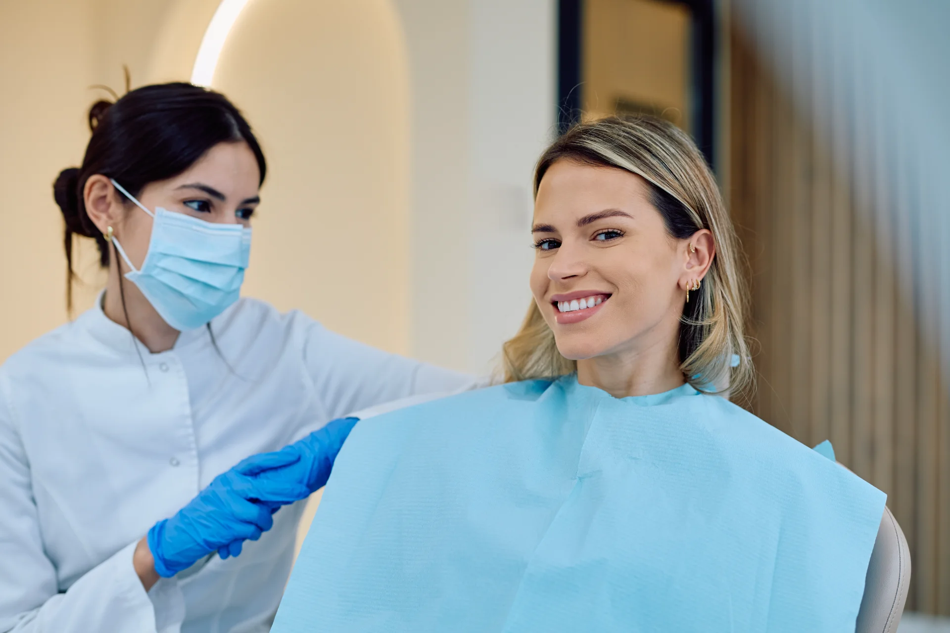 Female dentist in mask holding the hand of a smiling dental patient wearing a blue bib.