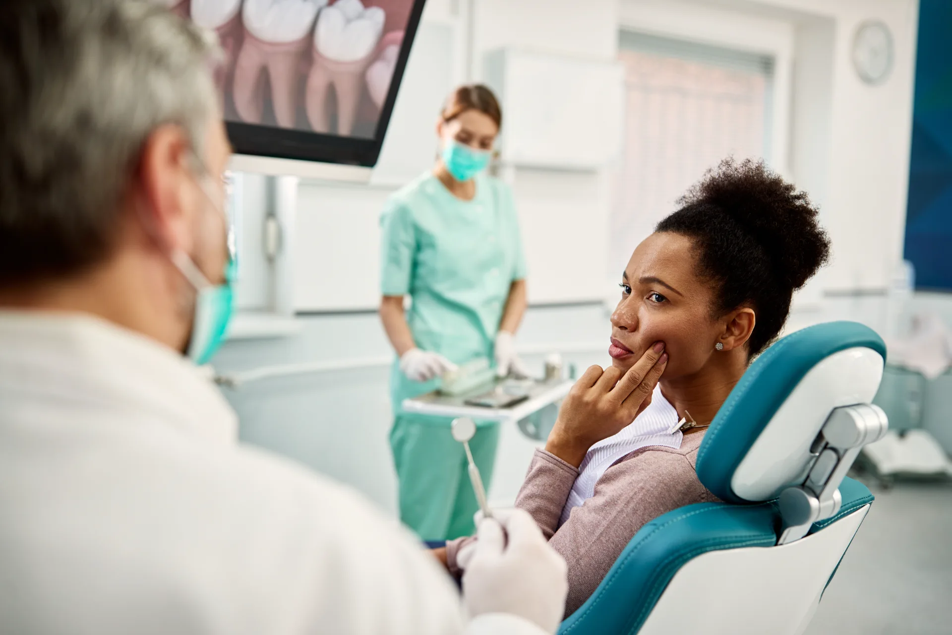 Woman in dental chair holds cheek in pain while dentist and assistant prepare tools in clinic.
