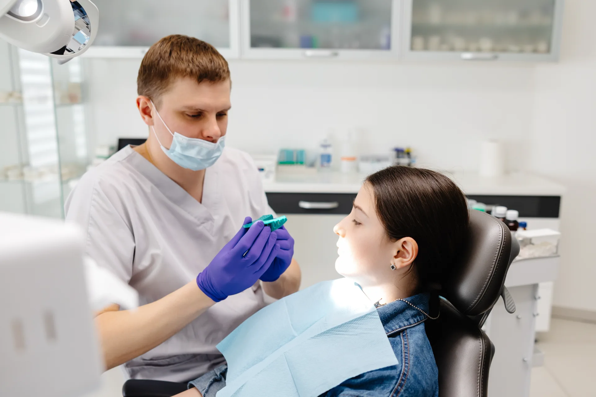 Dentist wearing gloves and mask showing a dental mold to a girl sitting in a dentist chair.