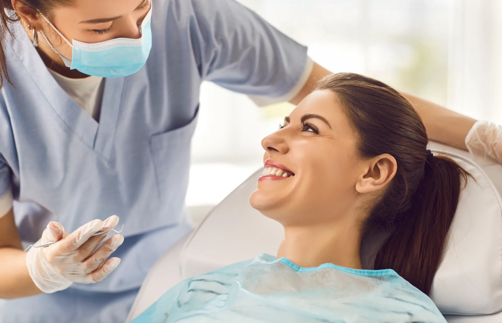 Female dentist with mask and gloves holds tool while smiling patient looks at her in dental chair.