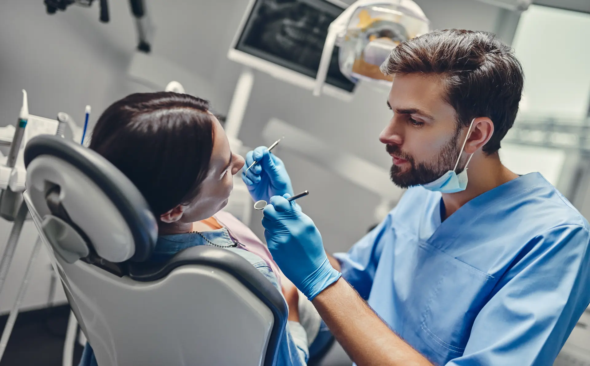 Dentist in blue scrubs examining a patient's teeth with dental tools in a clinic.