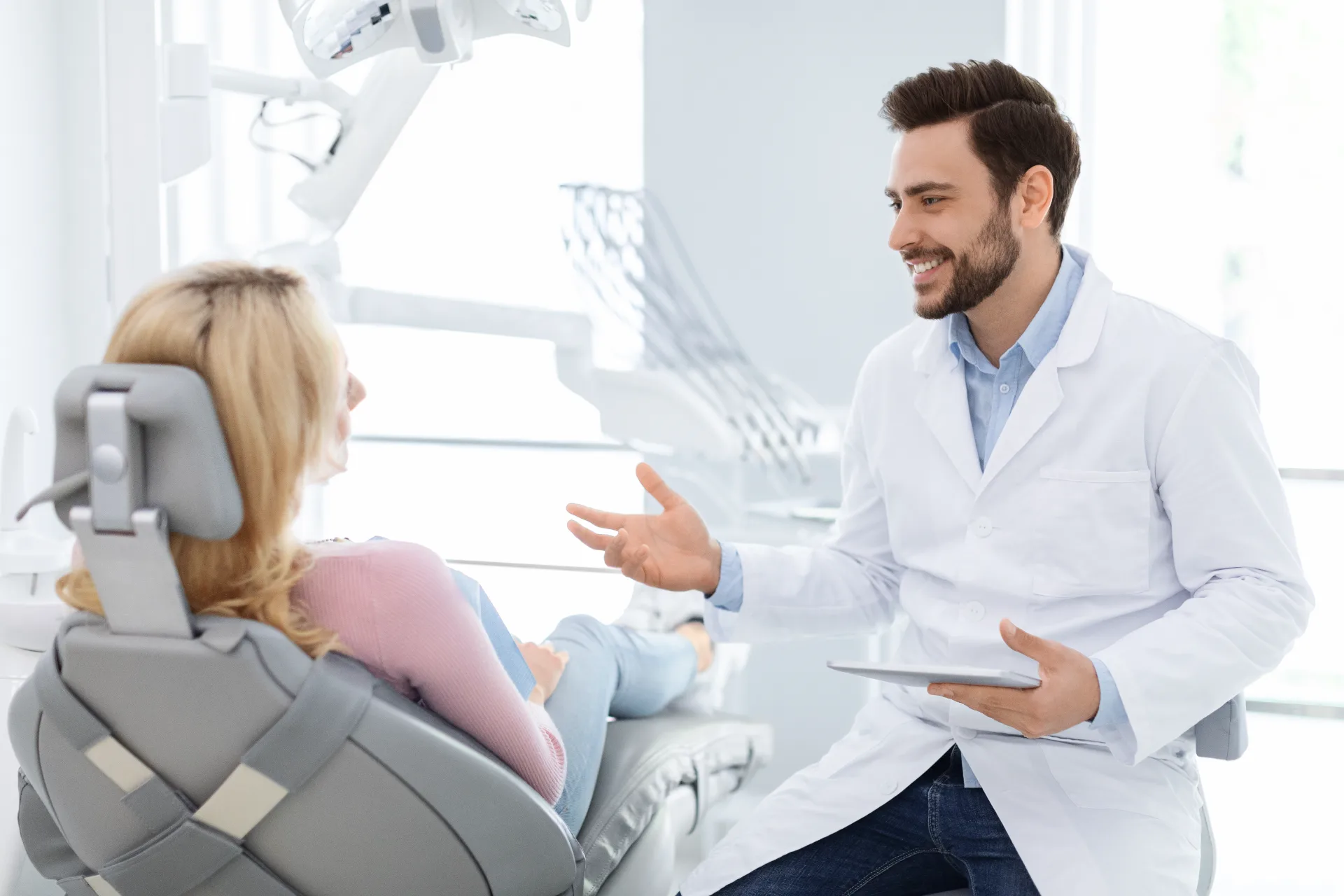 Dentist in white coat speaking with a seated female patient in a bright dental office.