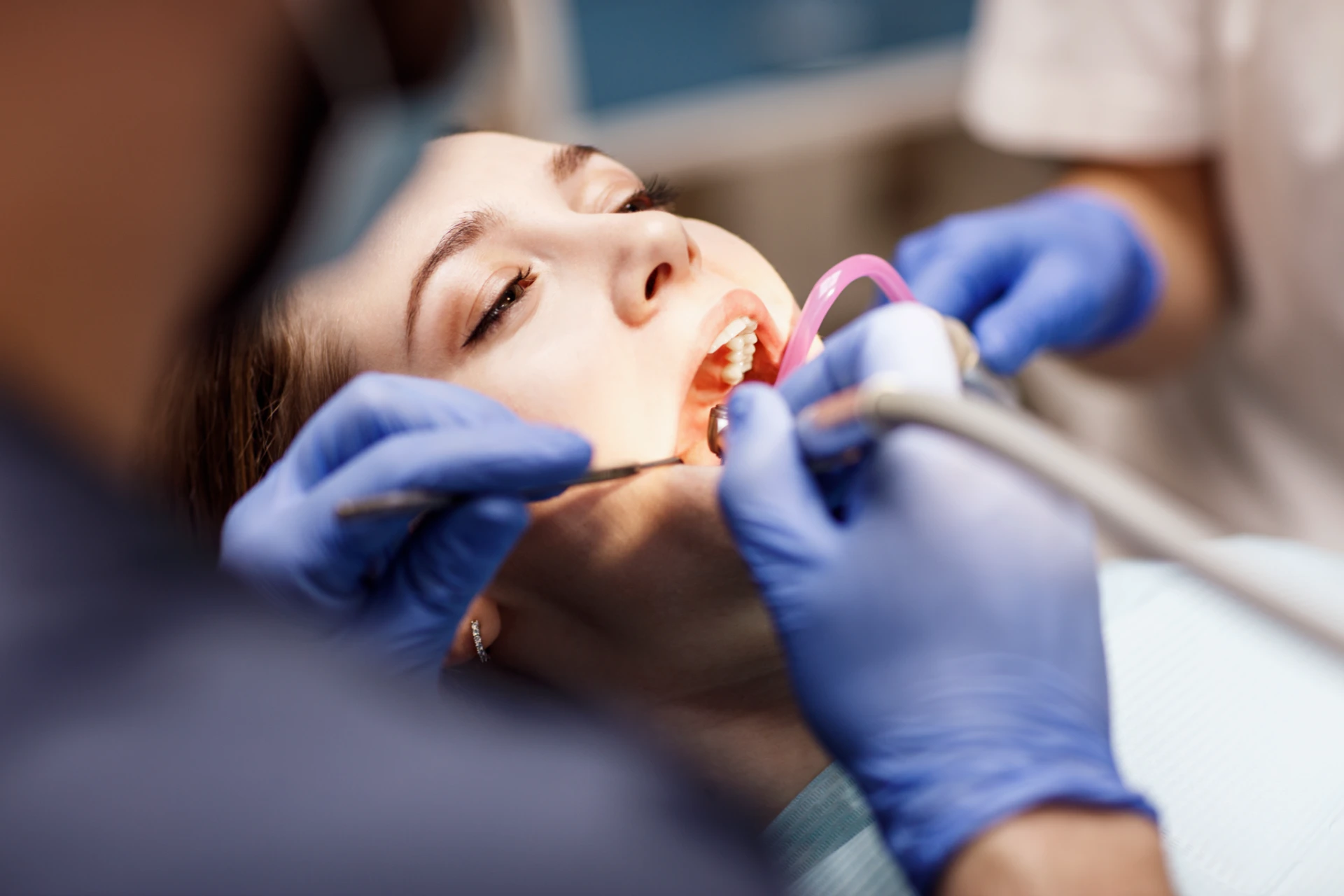 Dentist wearing blue gloves treating a female patient’s teeth with dental tools in her mouth.