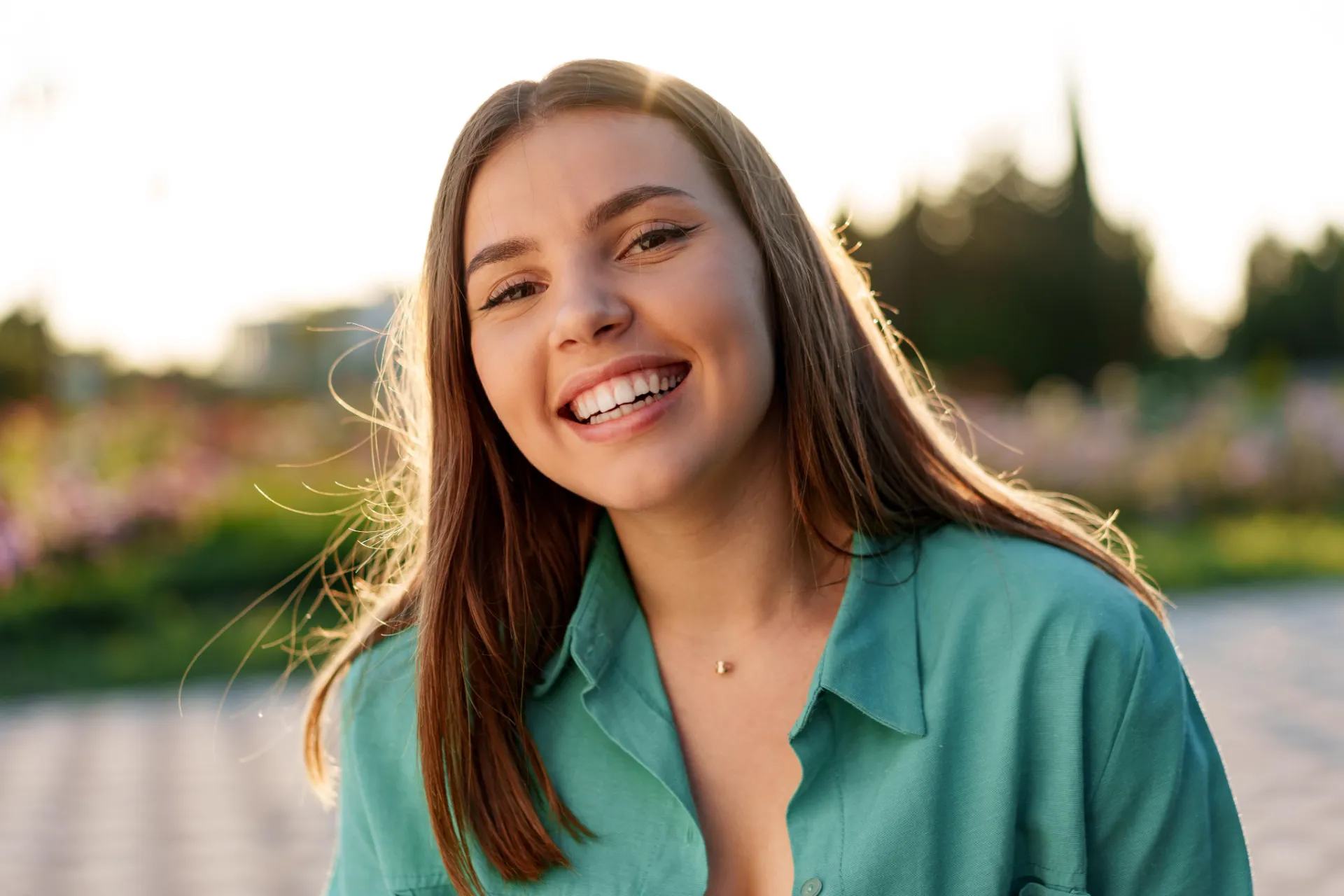 Smiling young woman with long hair wearing a teal shirt outdoors at sunset.