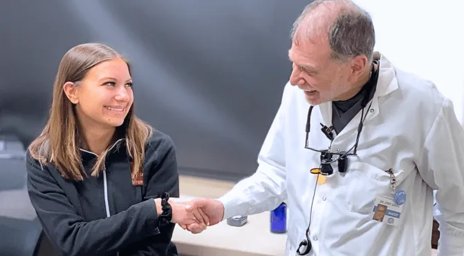 Smiling doctor in white coat shaking hands with a young woman in a black jacket indoors.