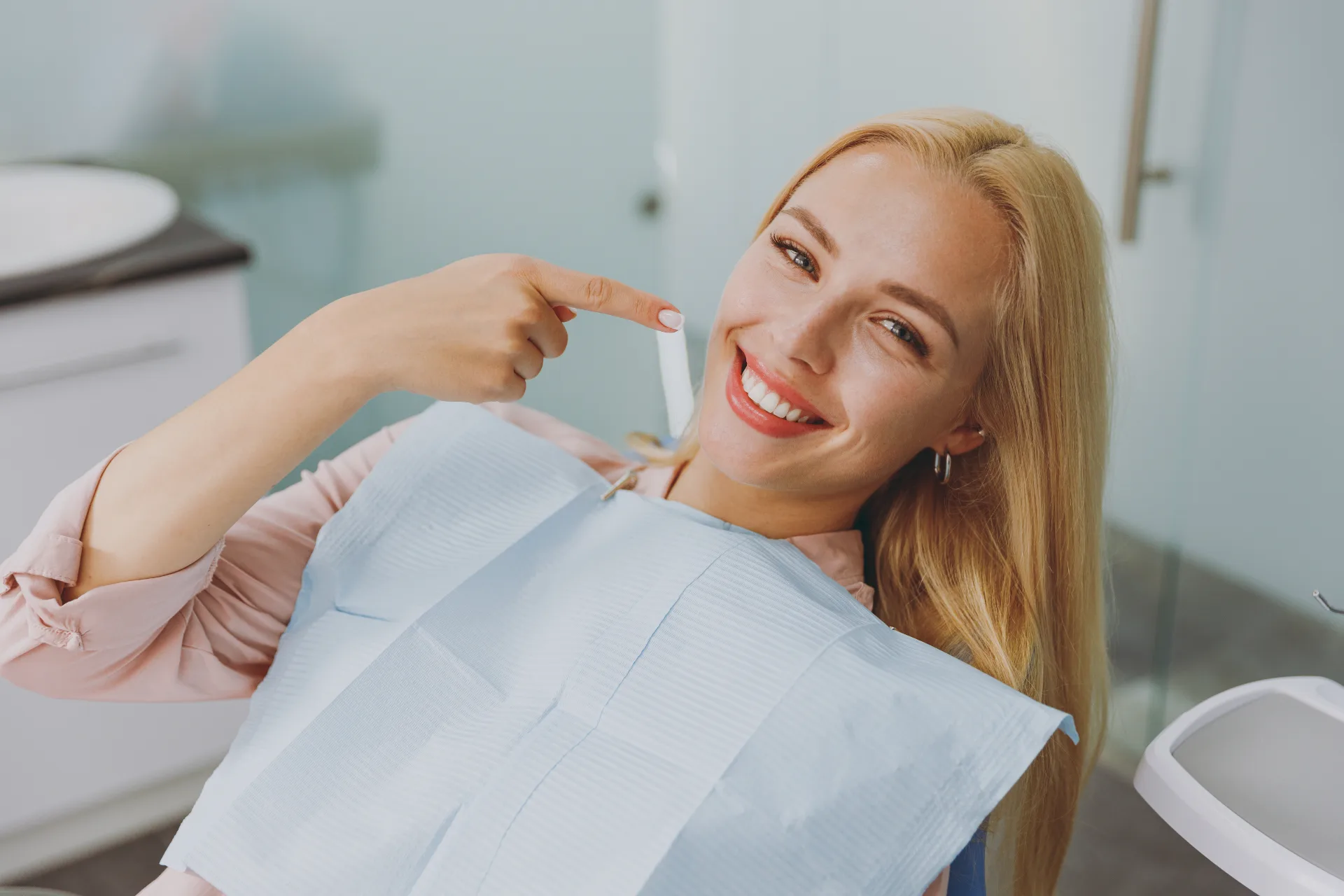 Smiling woman at dentist pointing to a bright white tooth in a dental clinic chair.