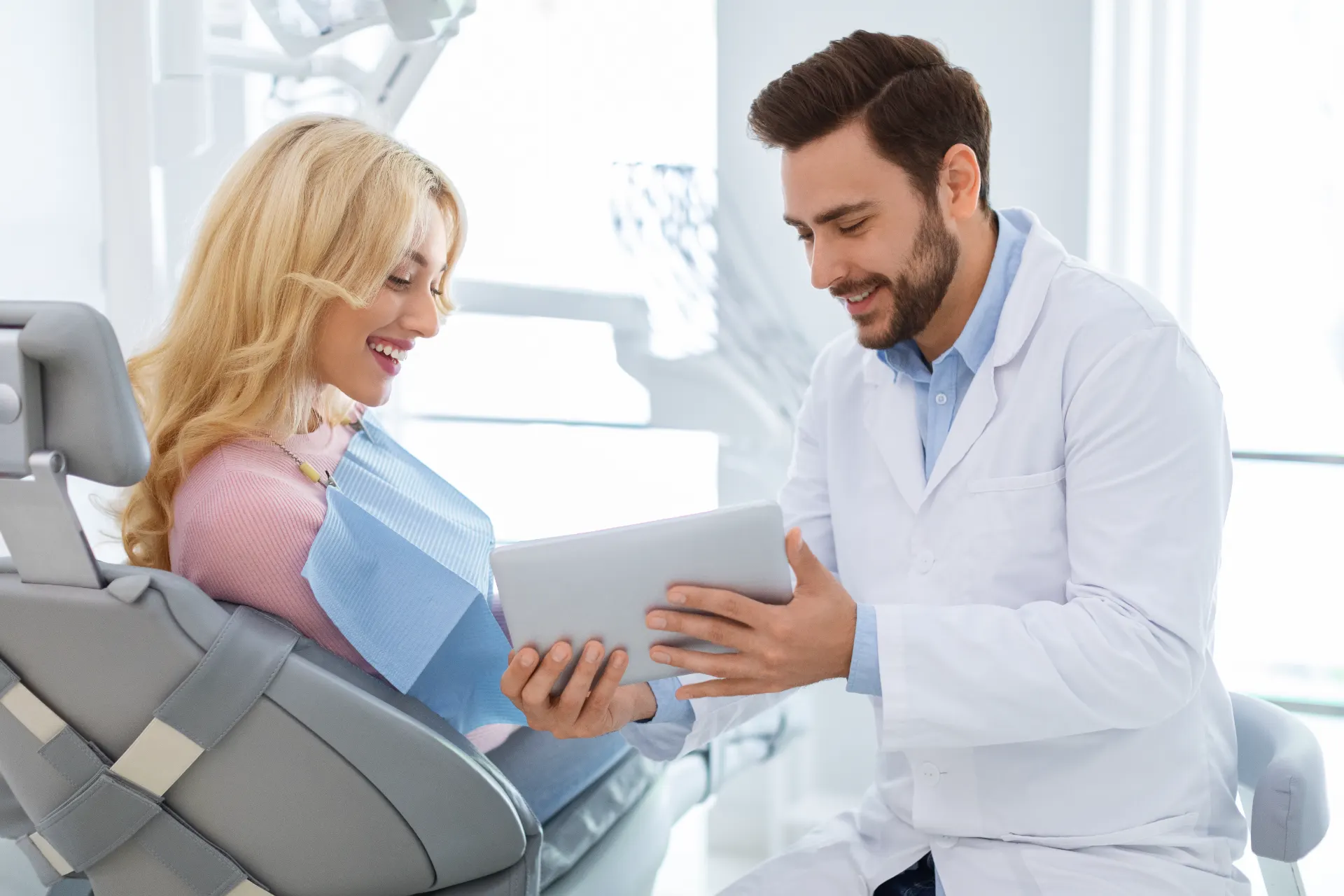 Dentist in white coat showing a smiling woman in dental chair something on a tablet.