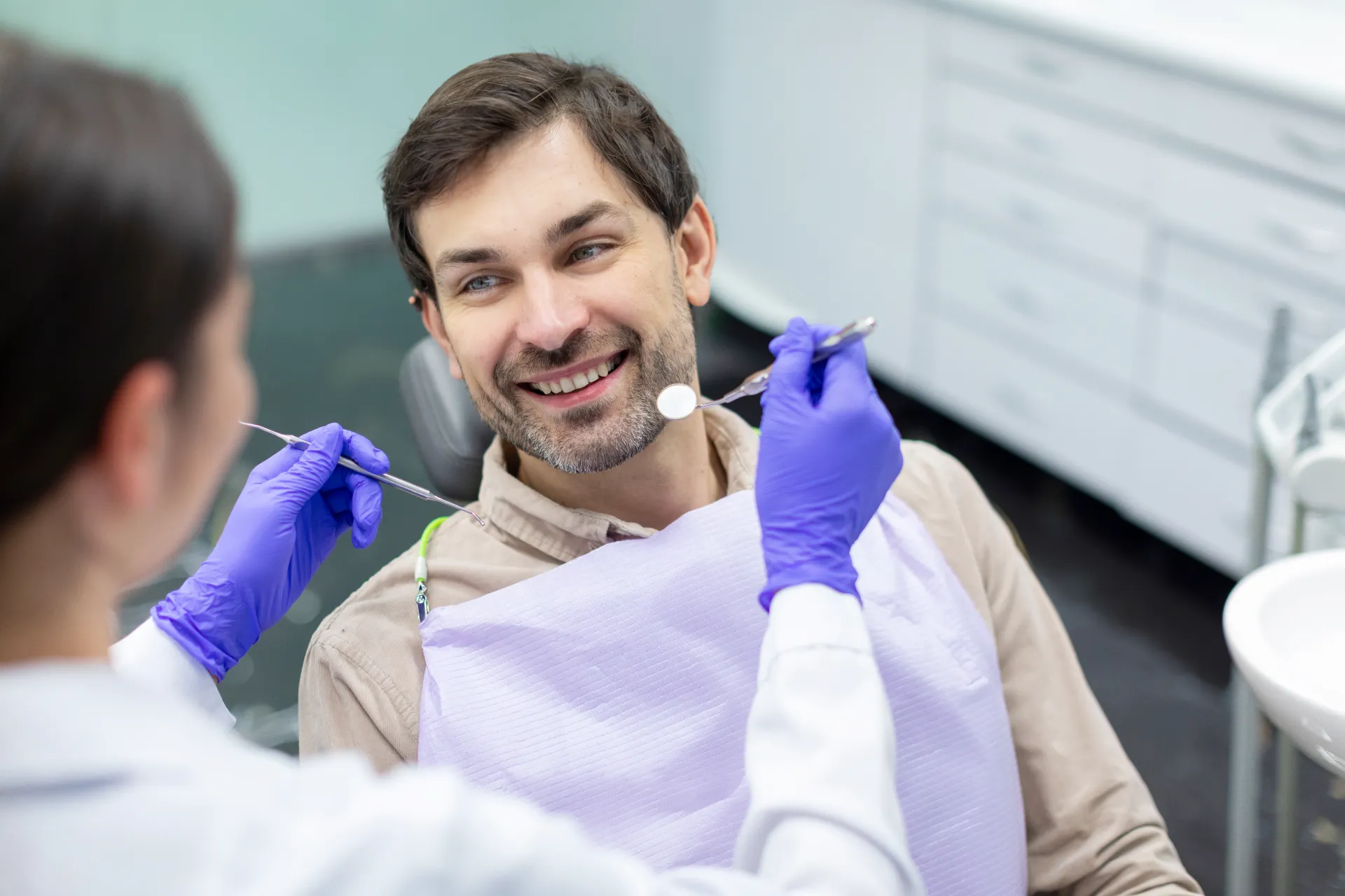 Smiling man in dental chair facing dentist holding dental mirror and tool wearing purple gloves.