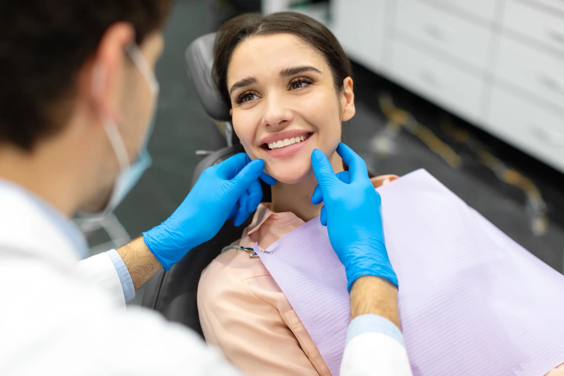 Dentist wearing blue gloves examines smiling woman's teeth at dental clinic.