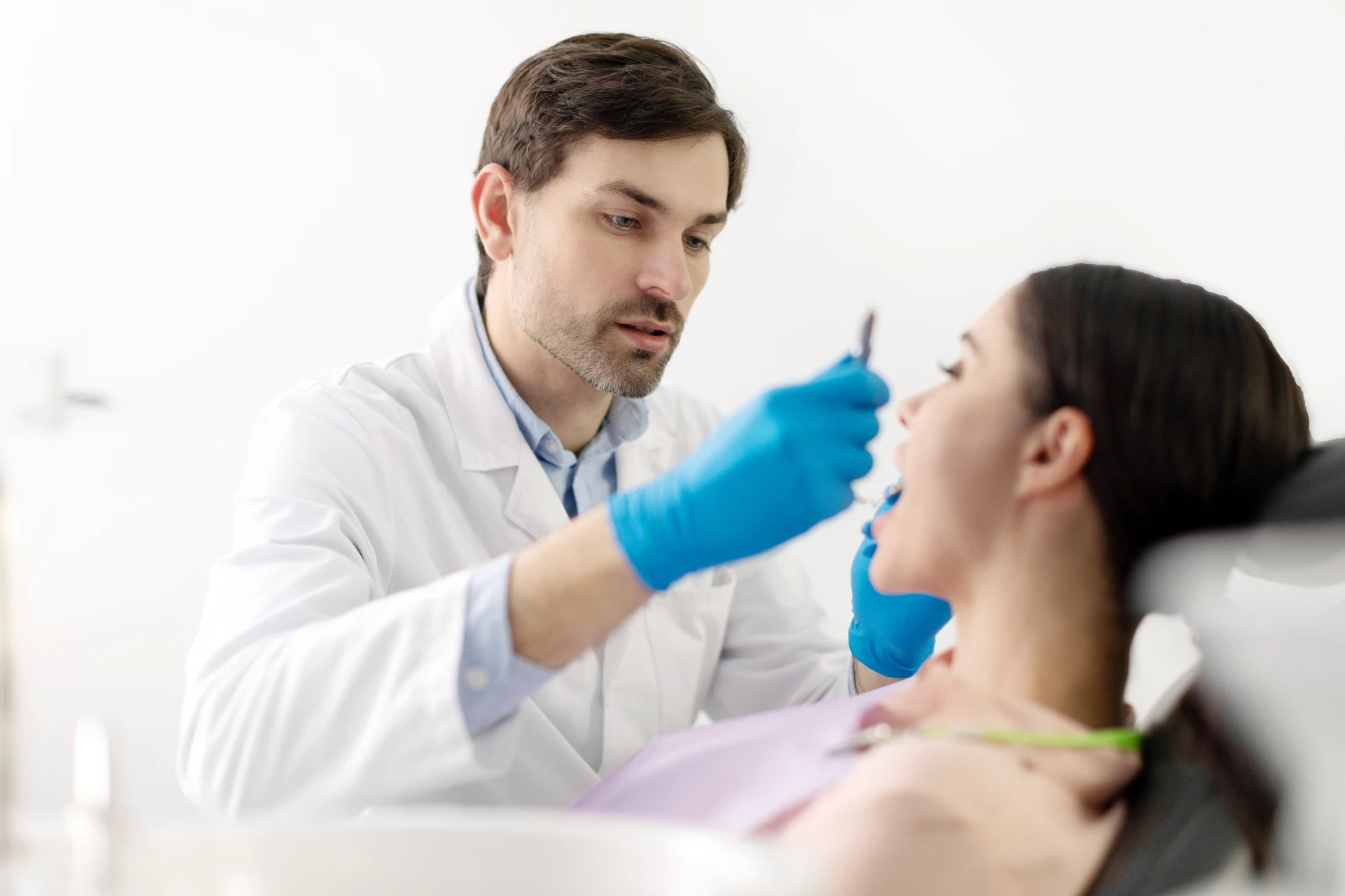 Male dentist wearing blue gloves examines a female patient's open mouth in a bright clinic.