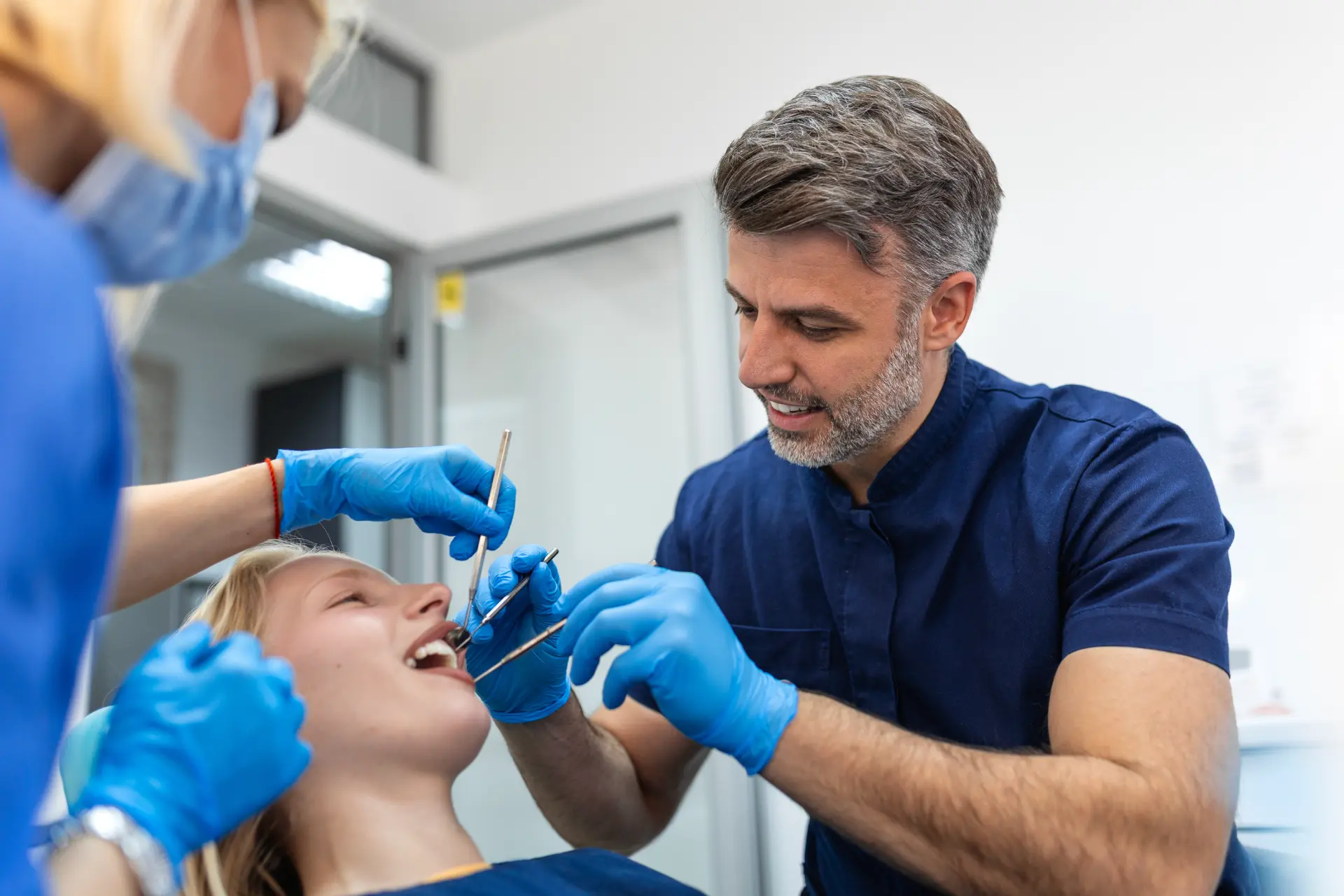 Dentist and assistant in blue gloves examining a female patient's teeth in clinic.