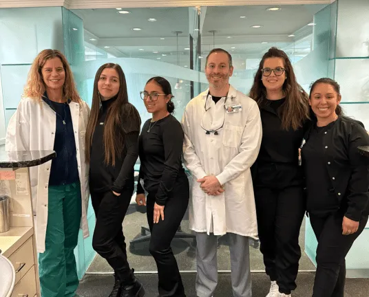 Group of six medical professionals standing together in a clinical setting wearing scrubs and lab coats.
