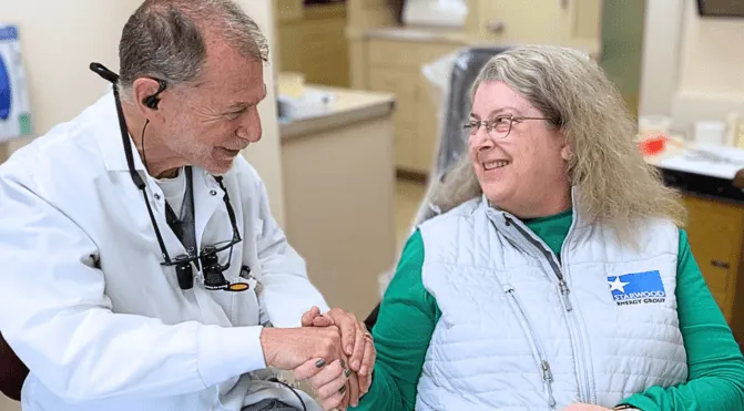 Older man in white coat holding hand of smiling middle-aged woman in a medical office.