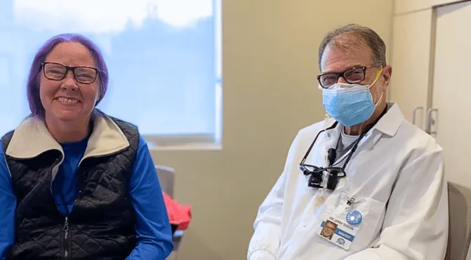 Smiling woman sits beside a doctor wearing a mask and white coat in a clinic room.