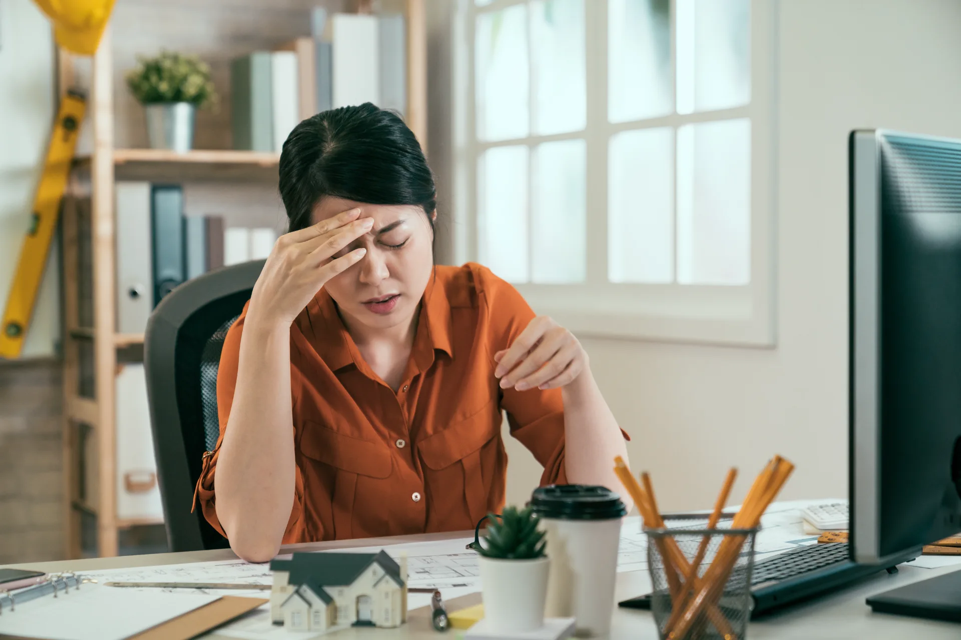 Woman in orange shirt sitting at desk with hand on forehead, appearing stressed or tired.