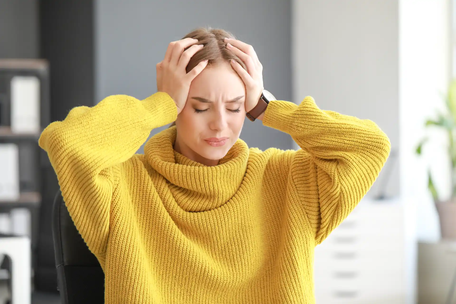 Woman in yellow sweater holding her head with eyes closed, appearing stressed or in pain at home.