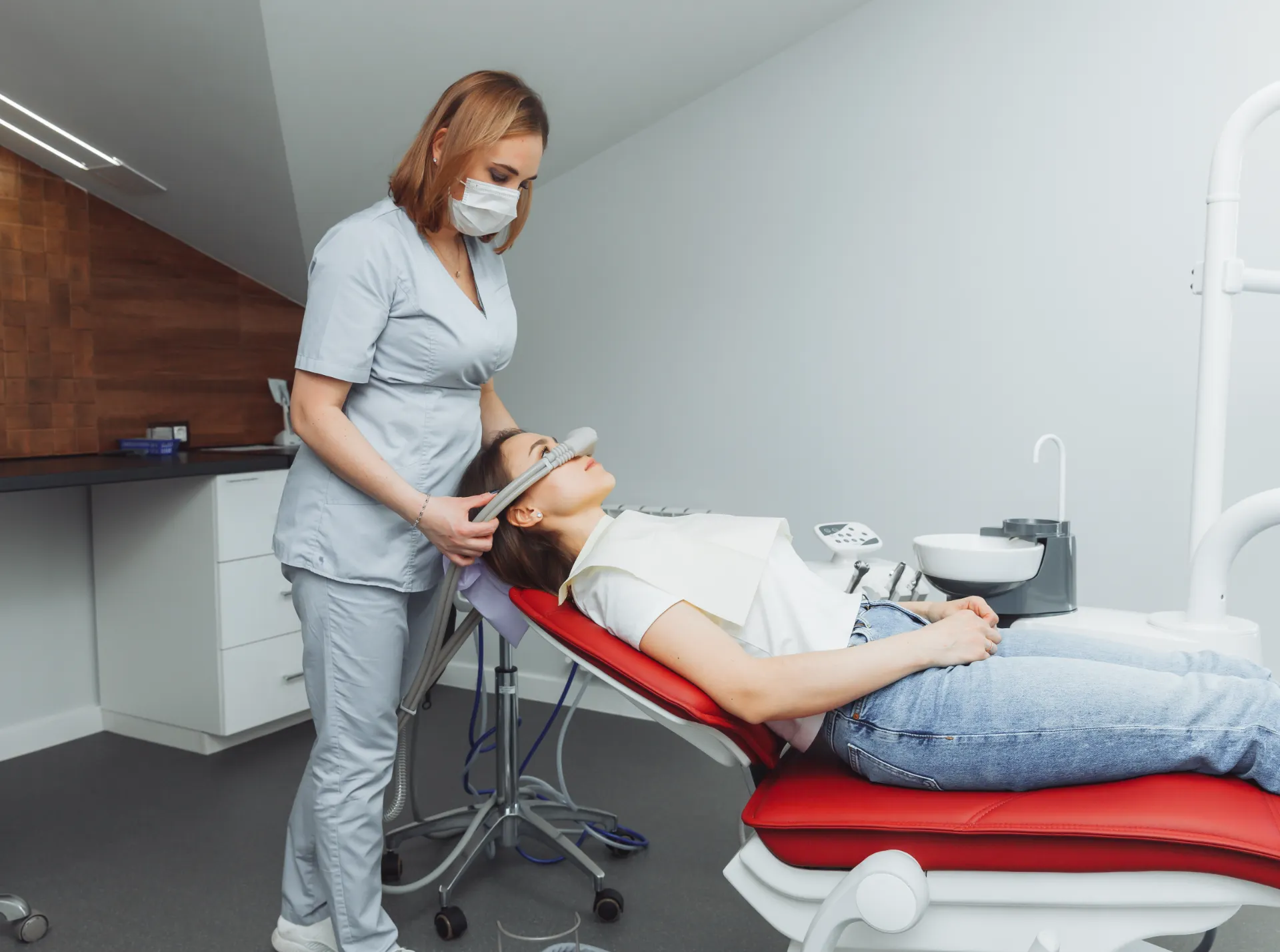 Dentist or hygienist in mask holding a tool near the nose of a patient lying on a red dental chair.