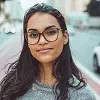 Young woman with long dark hair and glasses smiling outdoors on a city street.