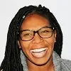 Smiling woman with glasses and long braided hair against a plain background.