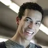 Smiling young man with short curly hair wearing a gray shirt in indoor lighting.