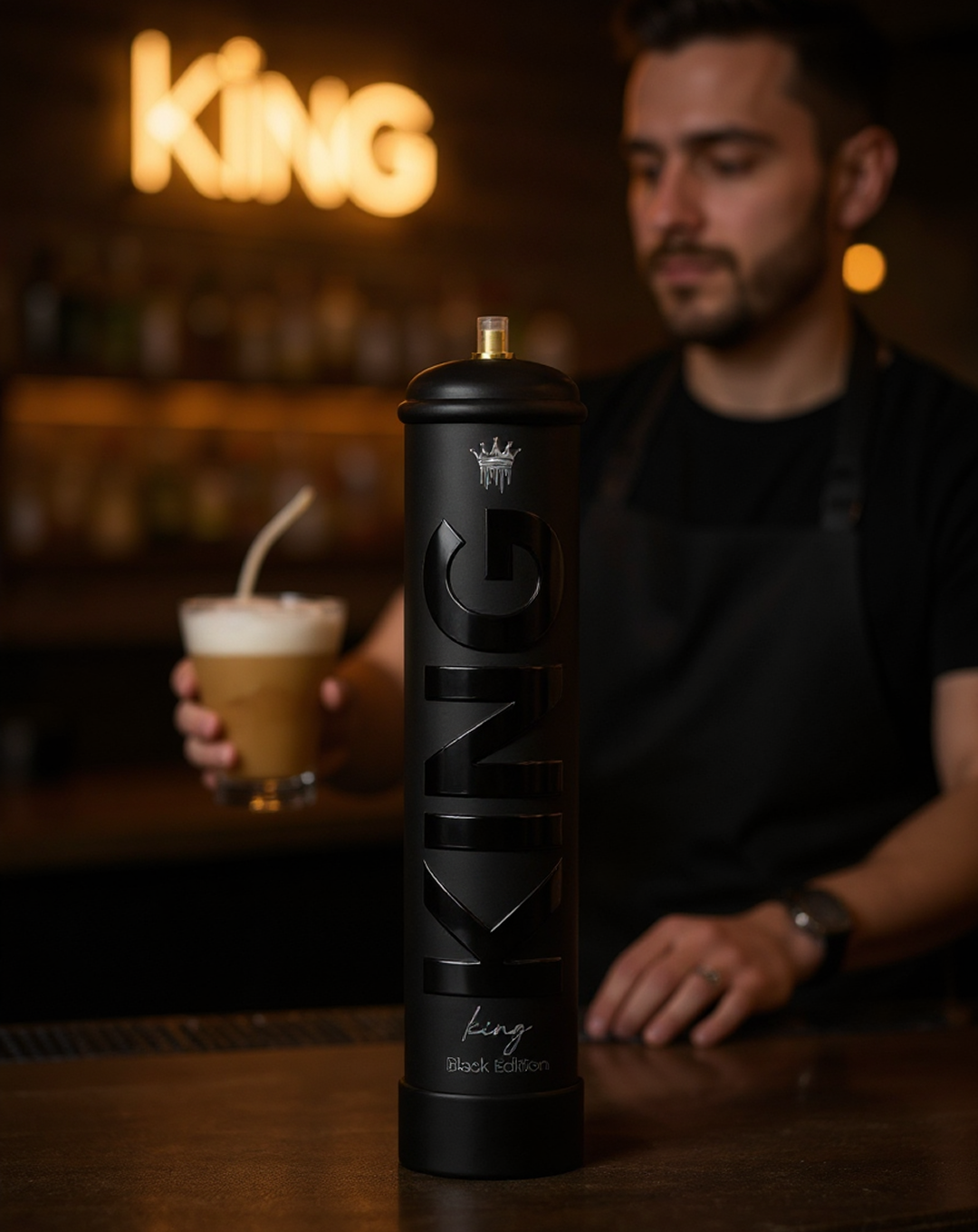 A tall black cylindrical container labeled 'KING Black Edition' with a crown symbol is centered on a bar counter, with a man in a black apron holding a glass of foamy coffee in the blurred background.