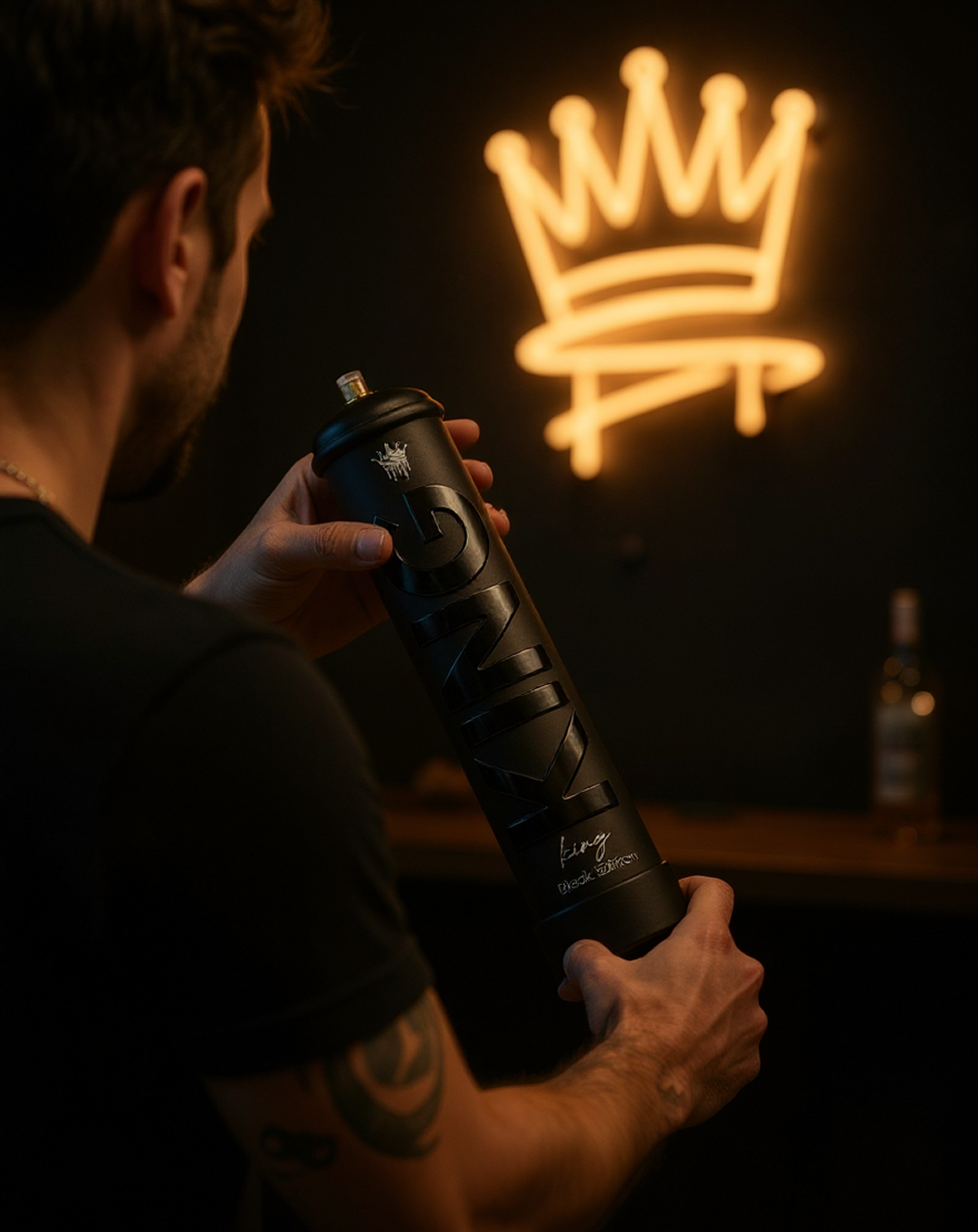 Man holding a matte black spray paint can with the word 'KING' in large letters on it and a small crown logo above, in a dimly lit room with a neon crown sign glowing in the background.