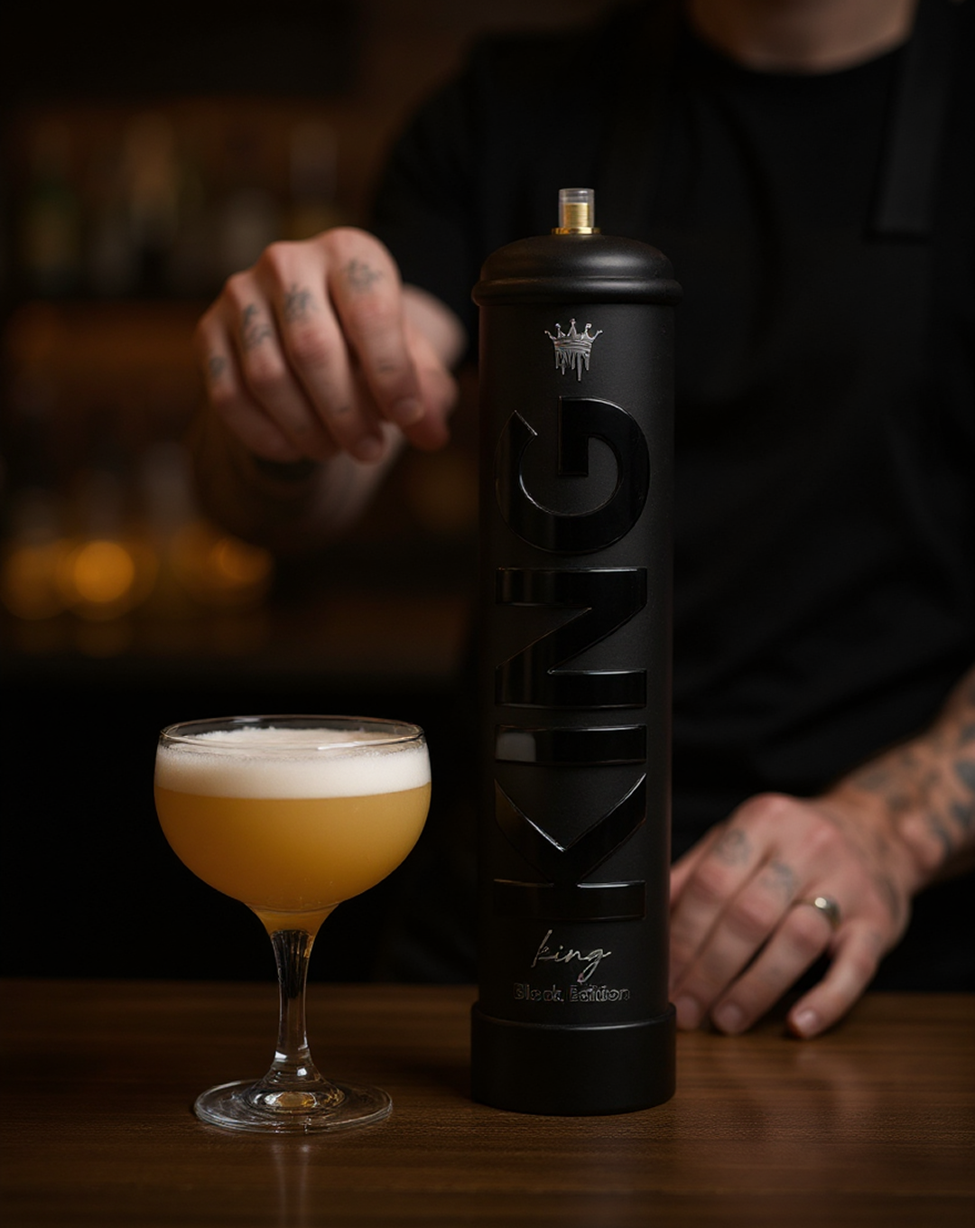 A person with tattooed hands and a black shirt pouring or preparing to pour a drink from a tall black container labeled 'KING Black Edition' next to a coupe glass filled with a foamy, amber-colored cocktail on a wooden surface.
