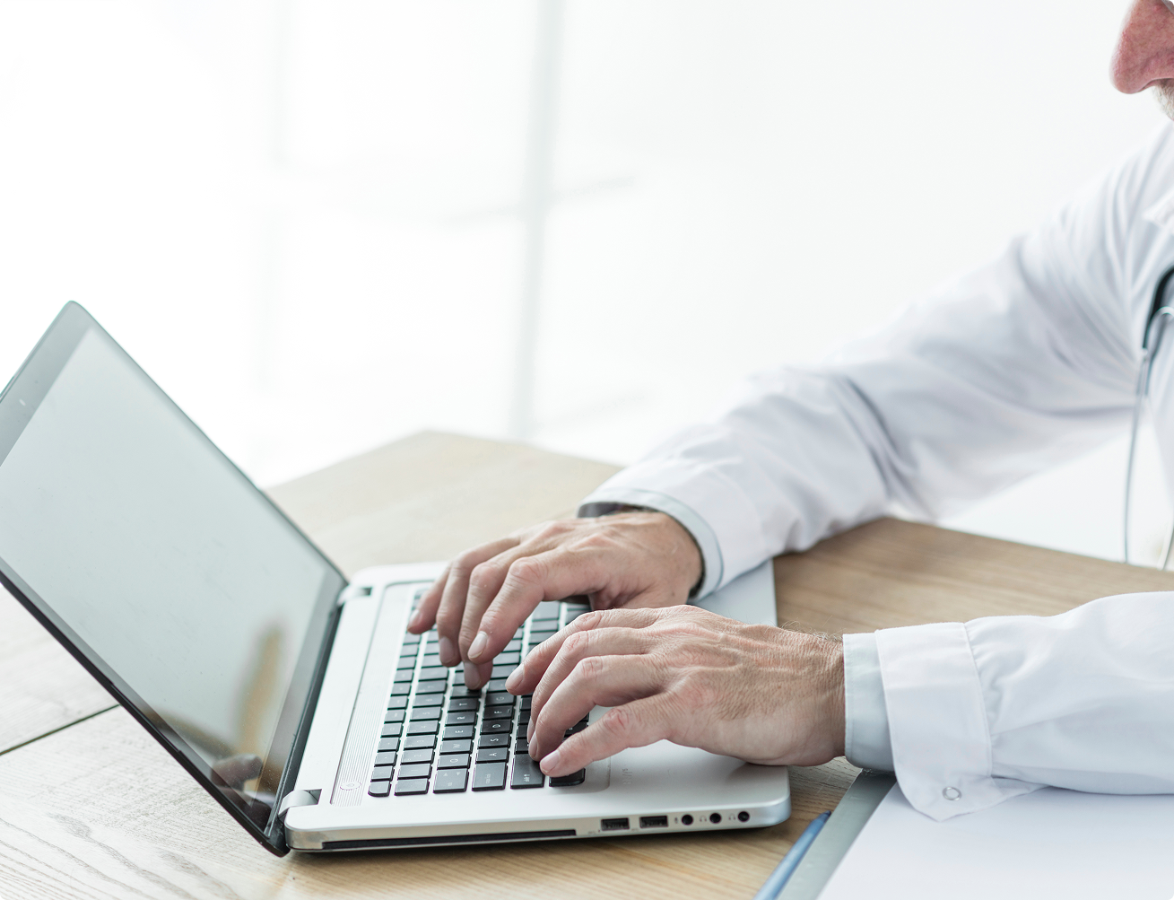 Person in white medical coat typing on a silver laptop on a wooden desk.