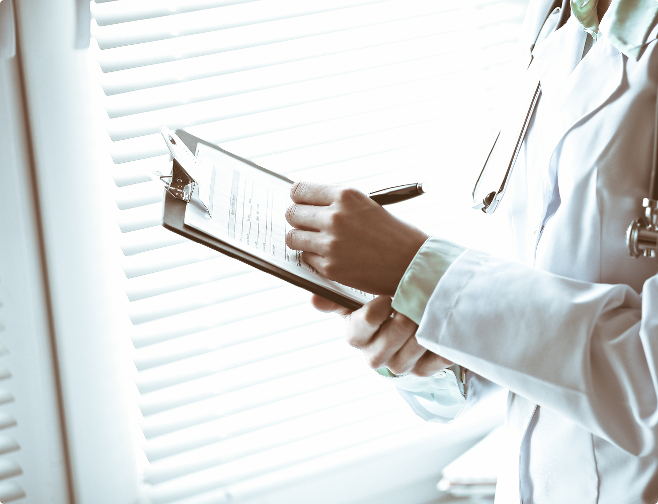 Doctor in white coat holding a clipboard and pen near window blinds.