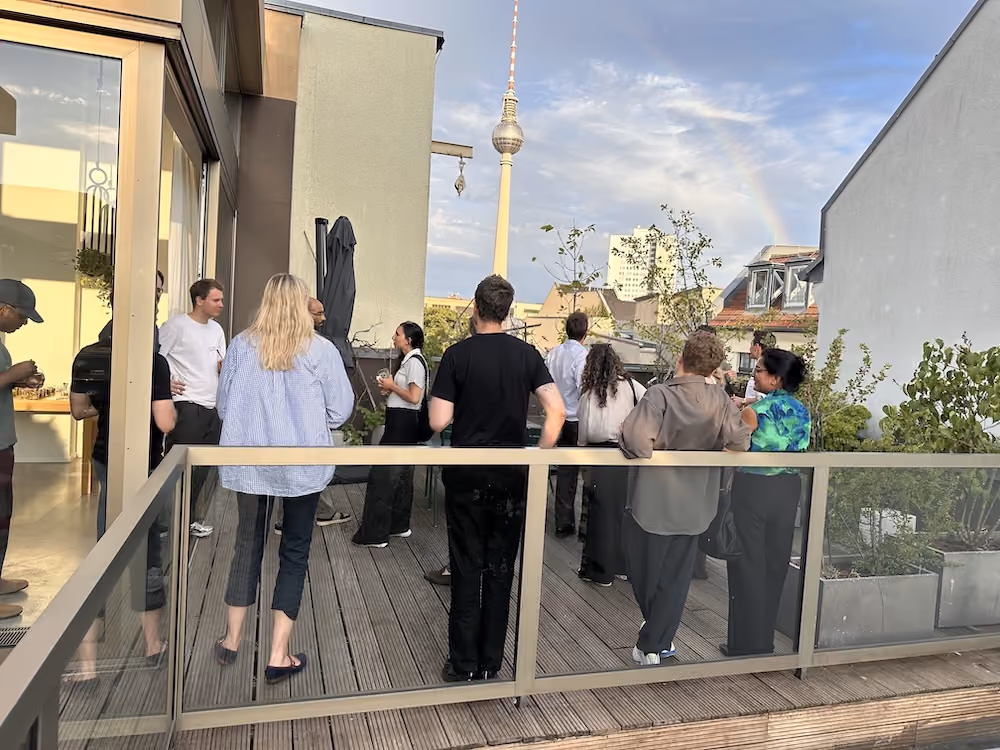 Group of people socializing on a rooftop terrace with TV tower and rainbow in background.