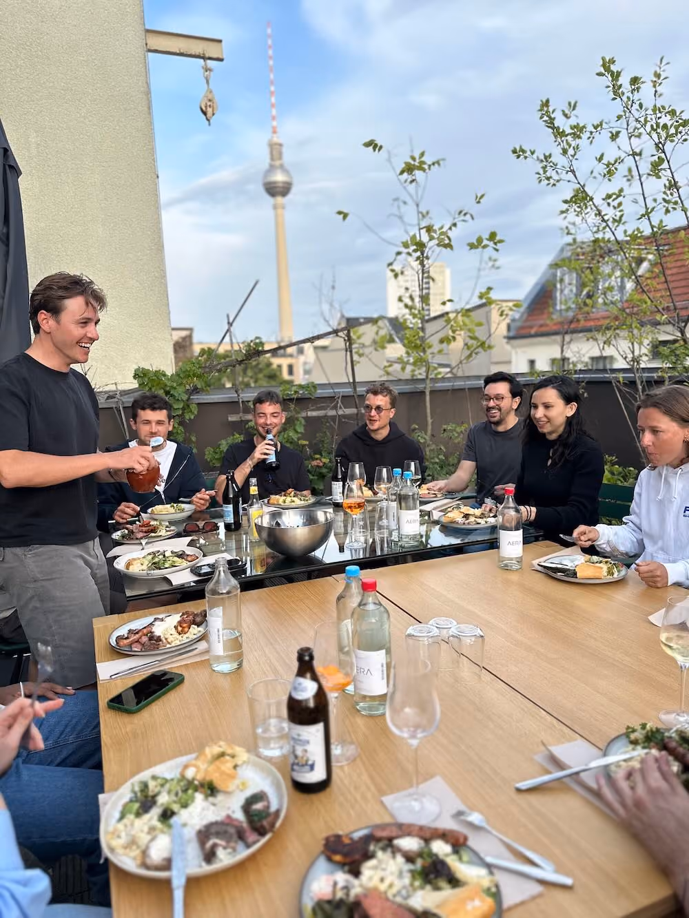 Group of people enjoying an outdoor meal on a rooftop with city view and beverages.