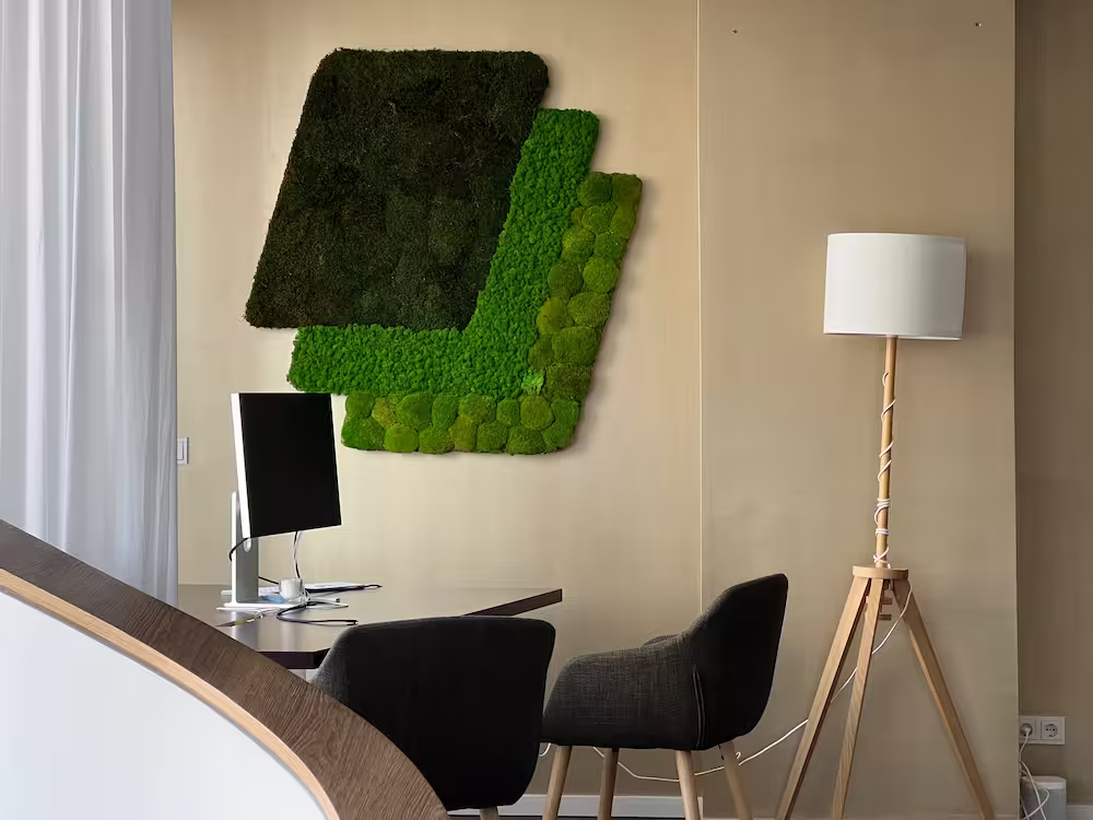 Modern office with black chairs, a computer on desk, green moss wall art, and a wooden tripod lamp.