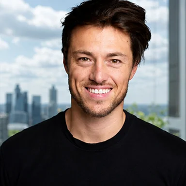 Smiling man with dark hair wearing black shirt in front of a window showing a city skyline.