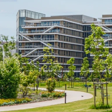 Modern multi-story building with balconies behind a landscaped park with trees and a walking path.