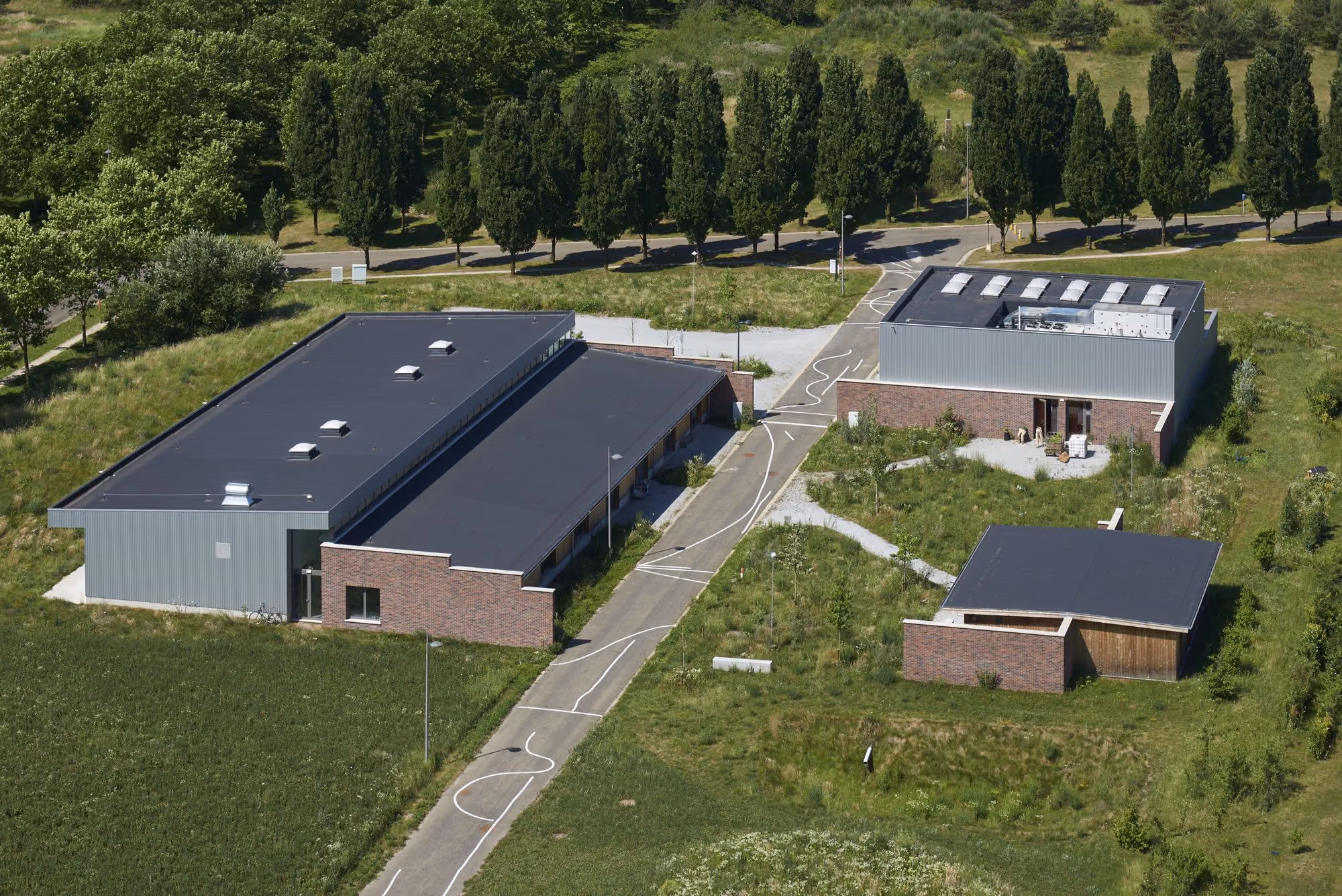Luchtfoto van drie moderne gebouwen met platte daken omgeven door gras en bomen.