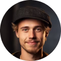 Smiling young man wearing a dark cap with a blurred dark background.