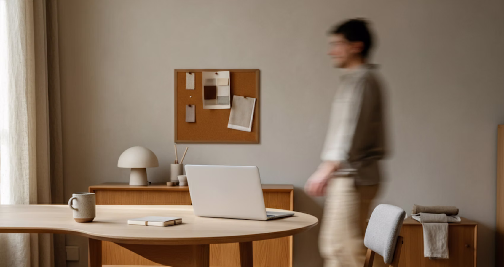 Minimalist workspace with a wooden desk, laptop, coffee mug, and blurred person walking by.