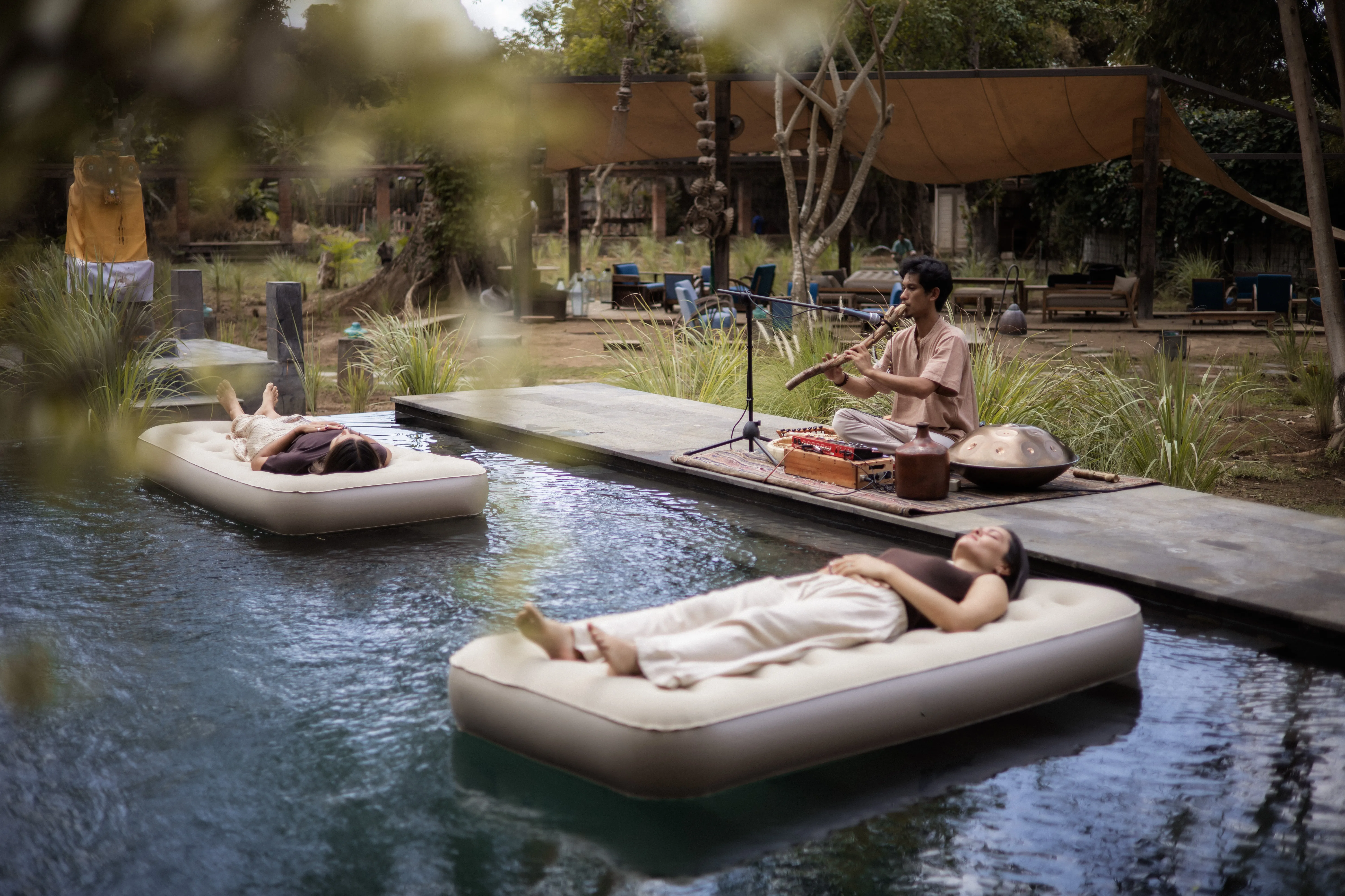 Retreat guests relaxing on air mattresses floating in a pool while a musician plays flute sound healing on the pool deck.