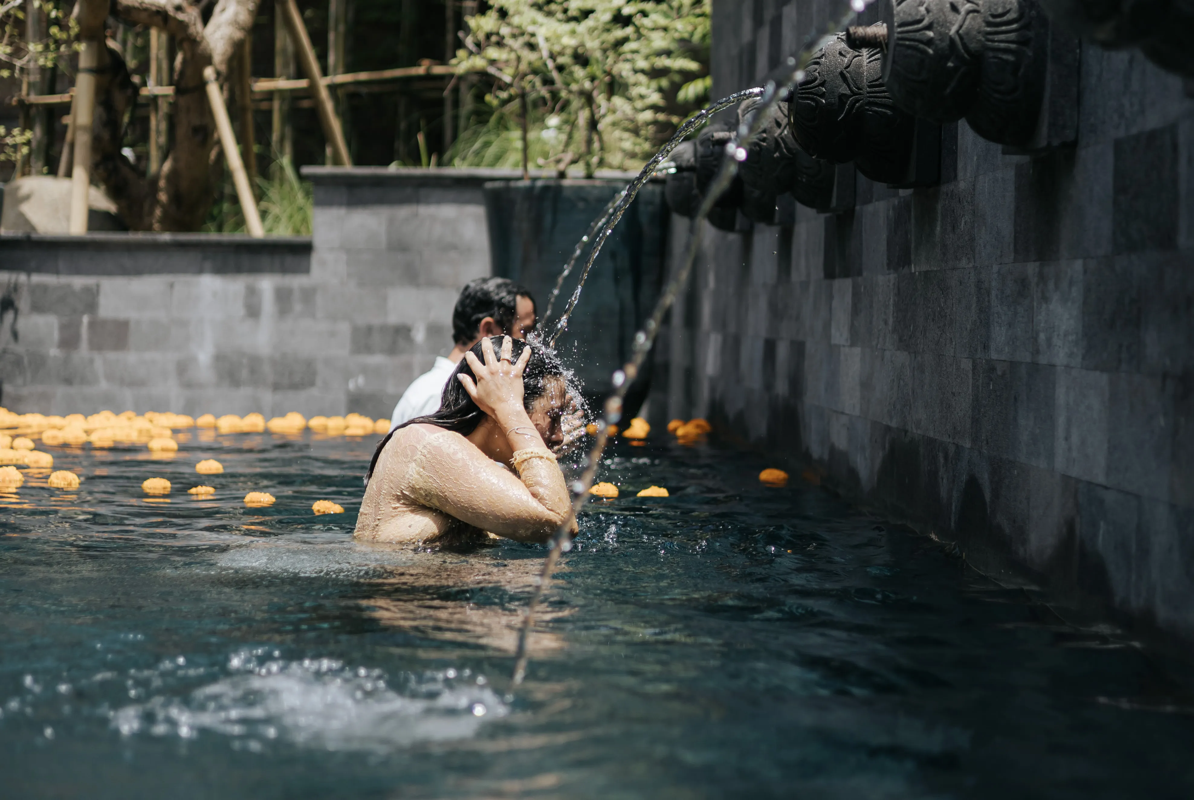 Woman undergoing a water cleansing ritual under a carved stone spout in a pool with floating marigold flowers at The Shaba Shala.