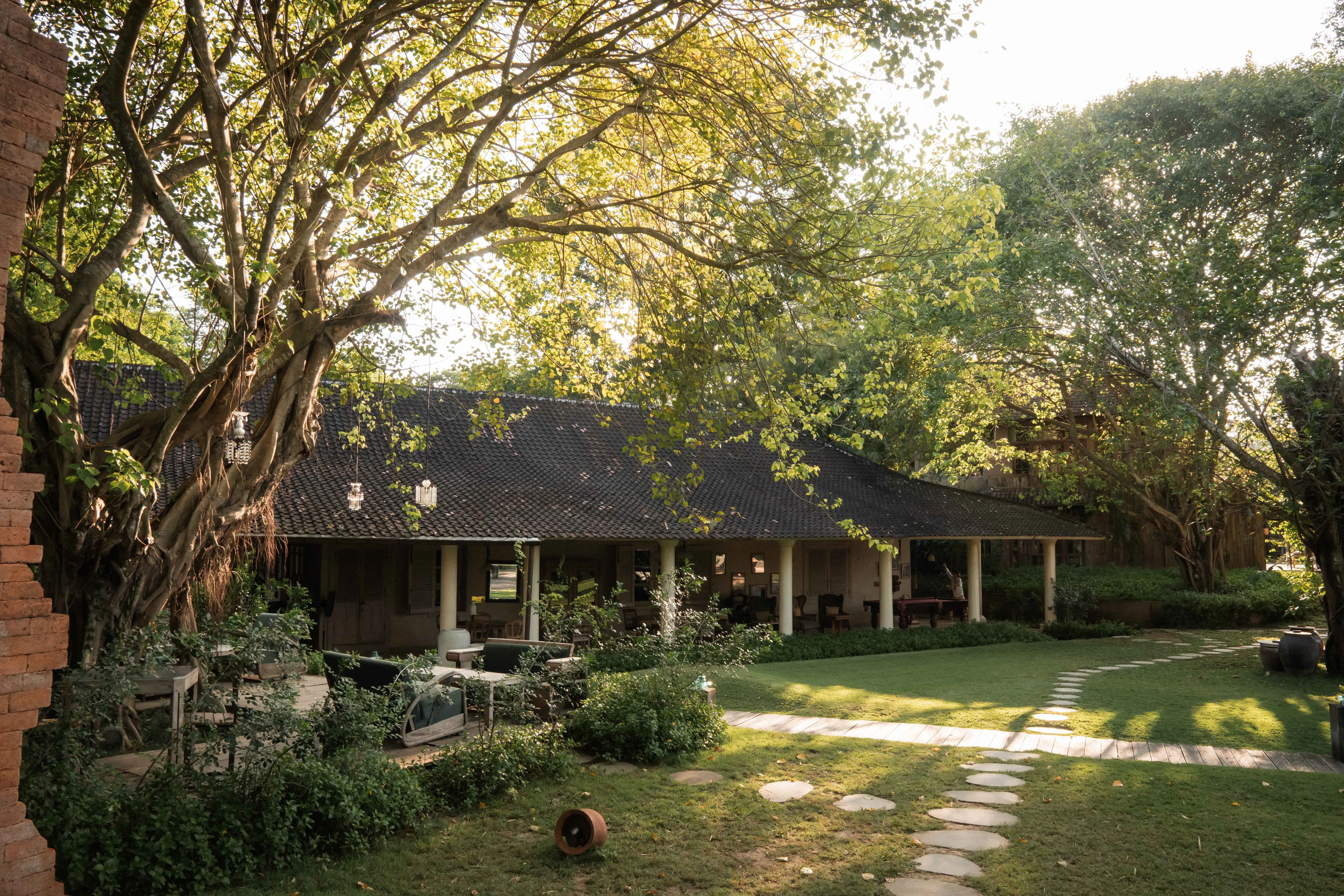 Large leafy trees shading a lawn and stepping-stone pathway leading to a traditional roofed pavilion at The Shaba Shala.