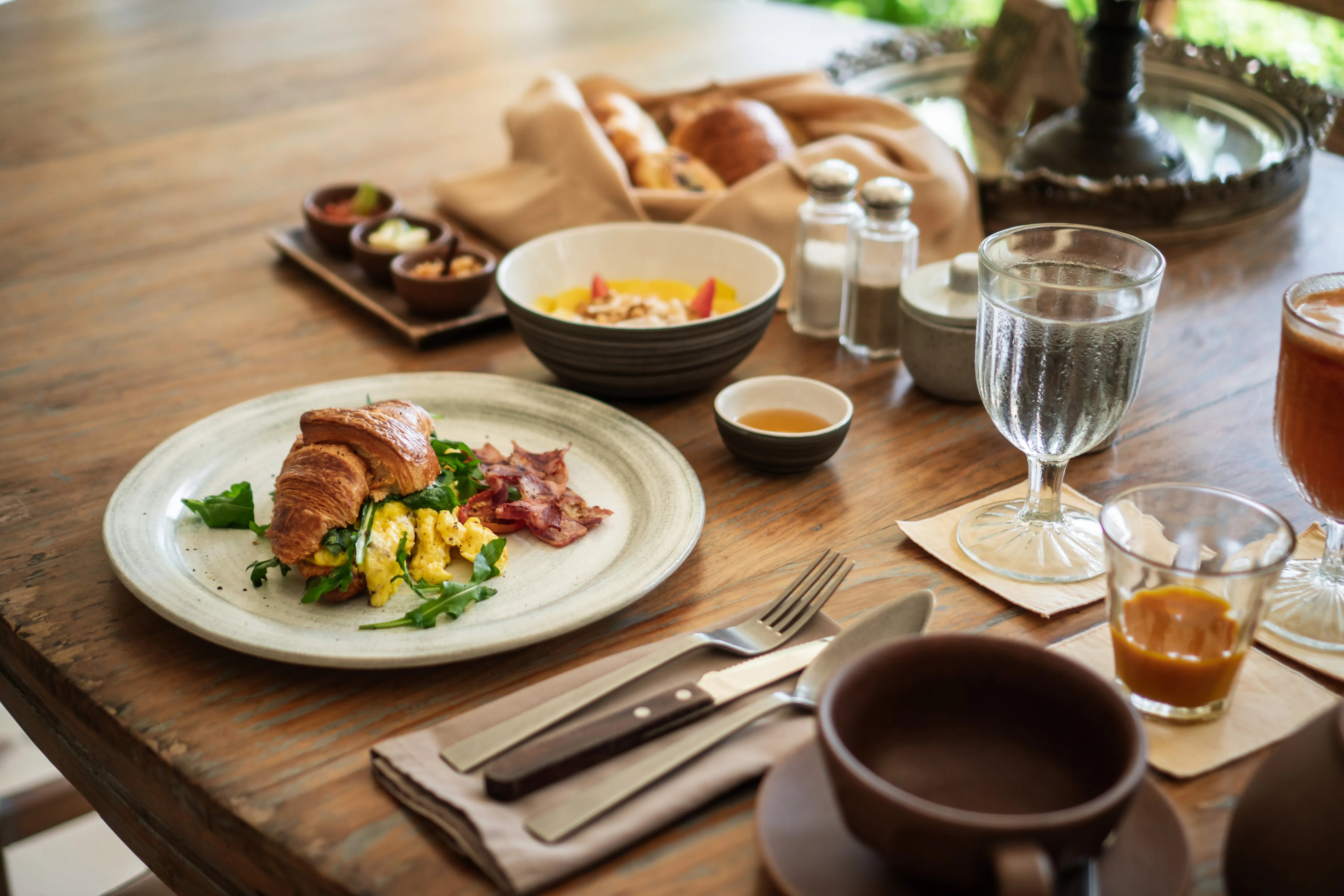 A freshly baked croissant sandwich with scrambled eggs and bacon served alongside fresh fruit and juices on a wooden table at The Shaba Shala.