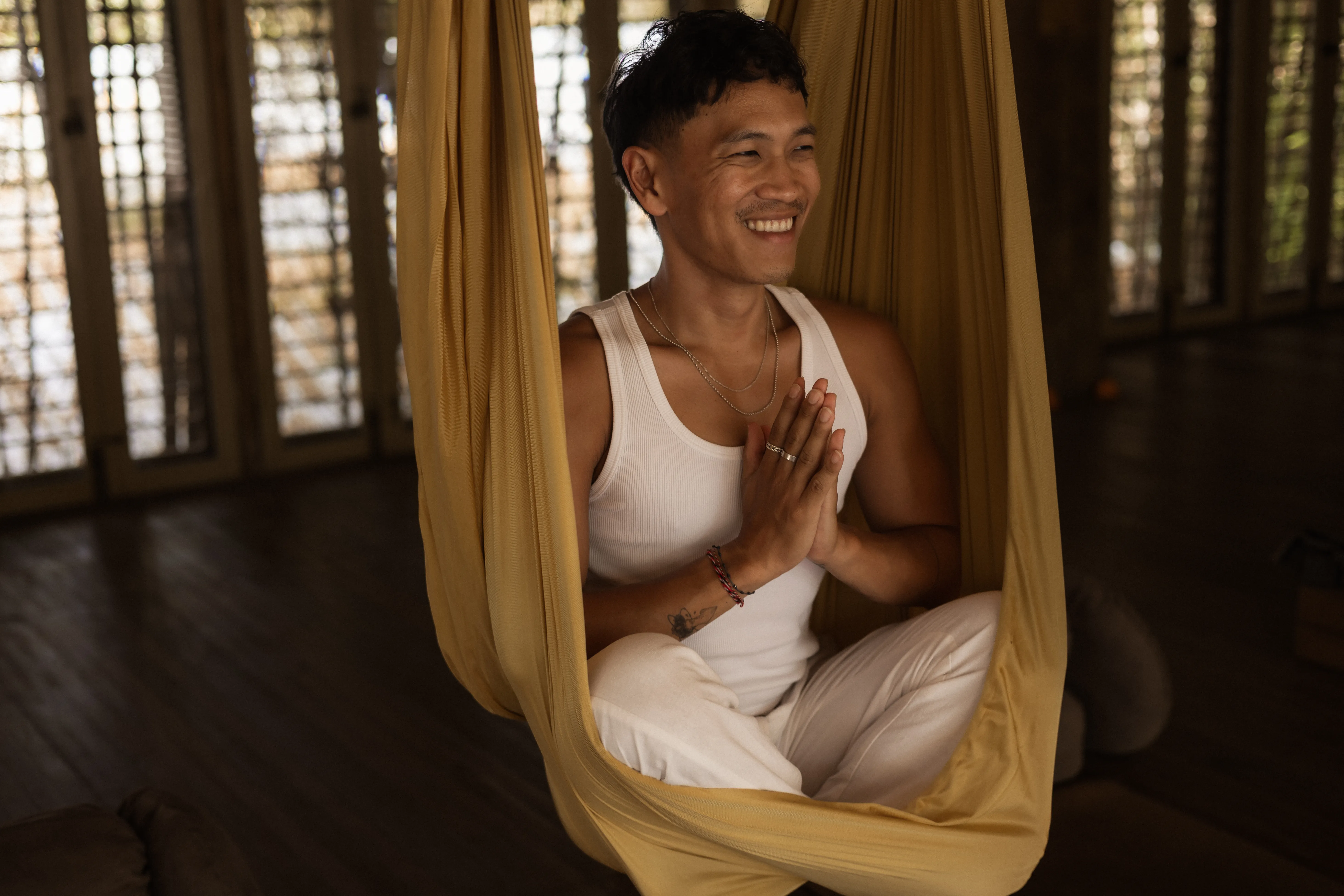 A man smiling with hands in prayer position while seated inside a yellow aerial yoga hammock at The Shaba Shala.
