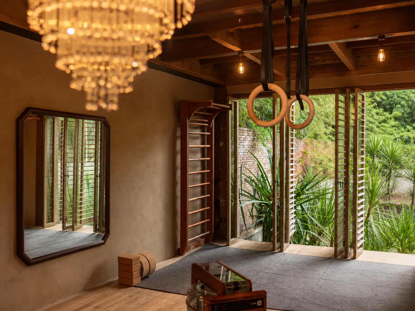 Boutique workout room with wooden gymnastics rings, wall bars, rowing machine, and louvered doors opening to greenery at The Shaba Shala.