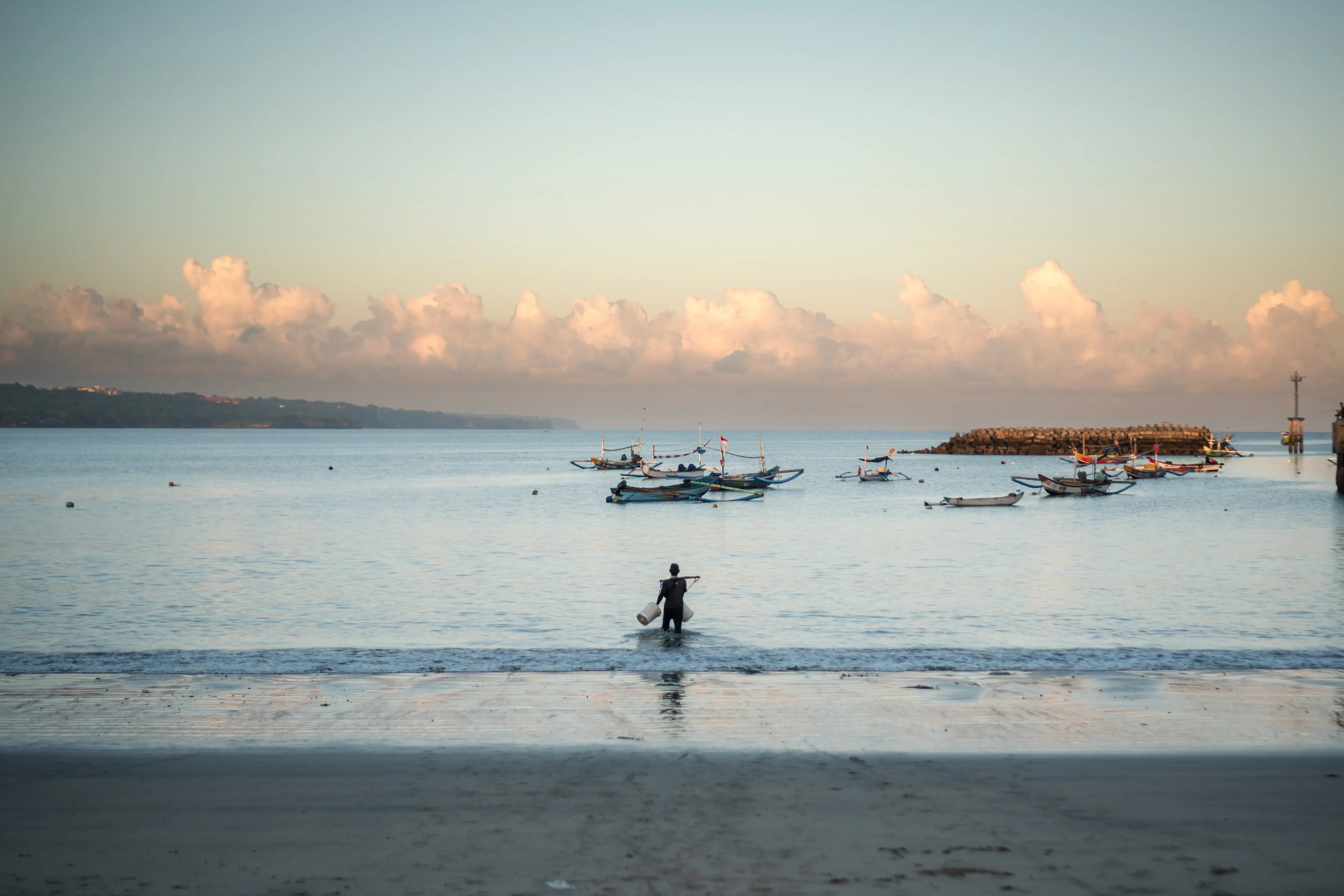 Calm ocean waters along a sandy beach in Bali with traditional fishing boats floating near the horizon during early morning light.