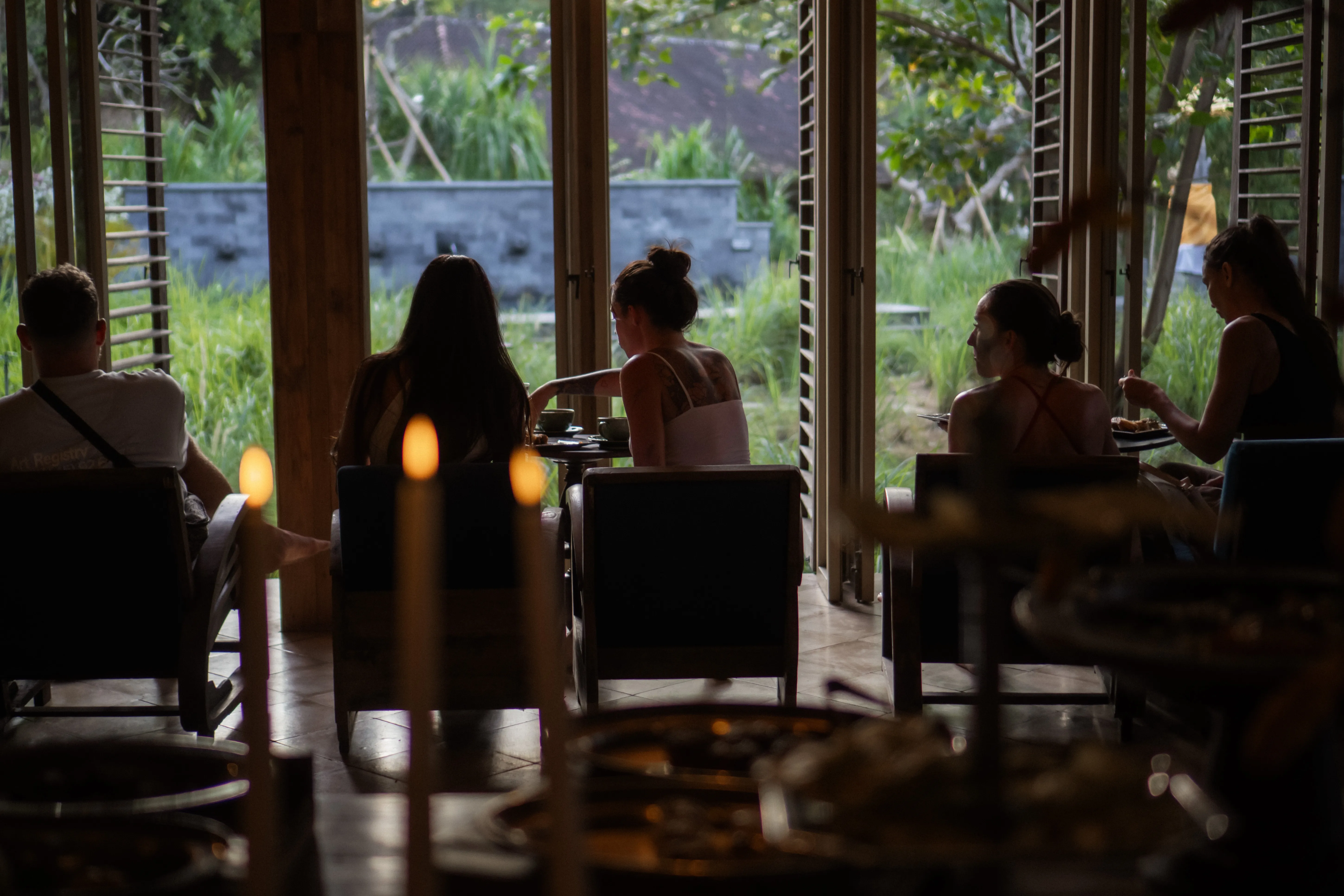 Silhouetted retreat guests seated in comfortable armchairs enjoying a meal by open sliding doors with warm candlelight in the foreground.