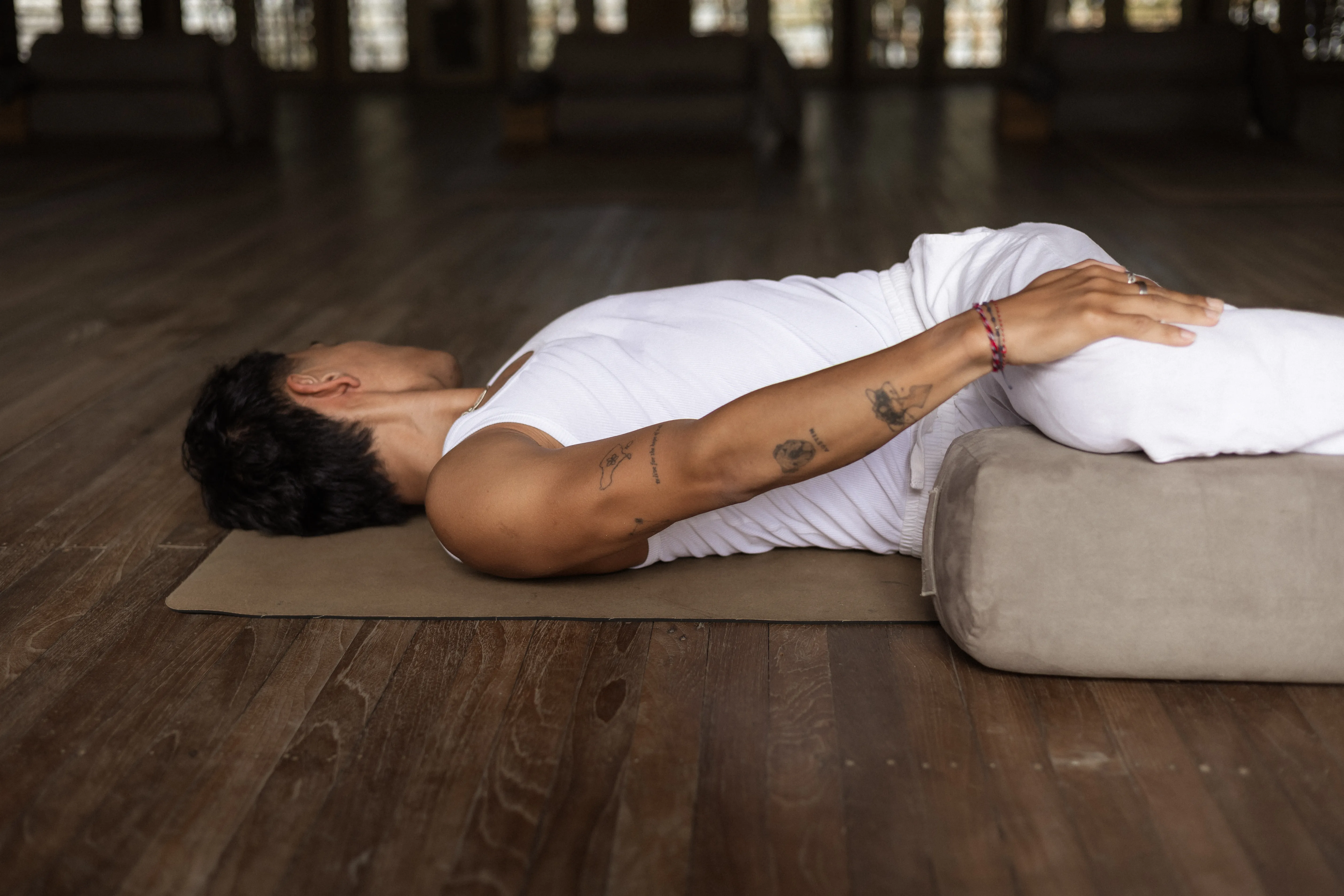Man dressed in white relaxing on a yoga mat with legs supported on a grey bolster at The Shaba Shala studio.
