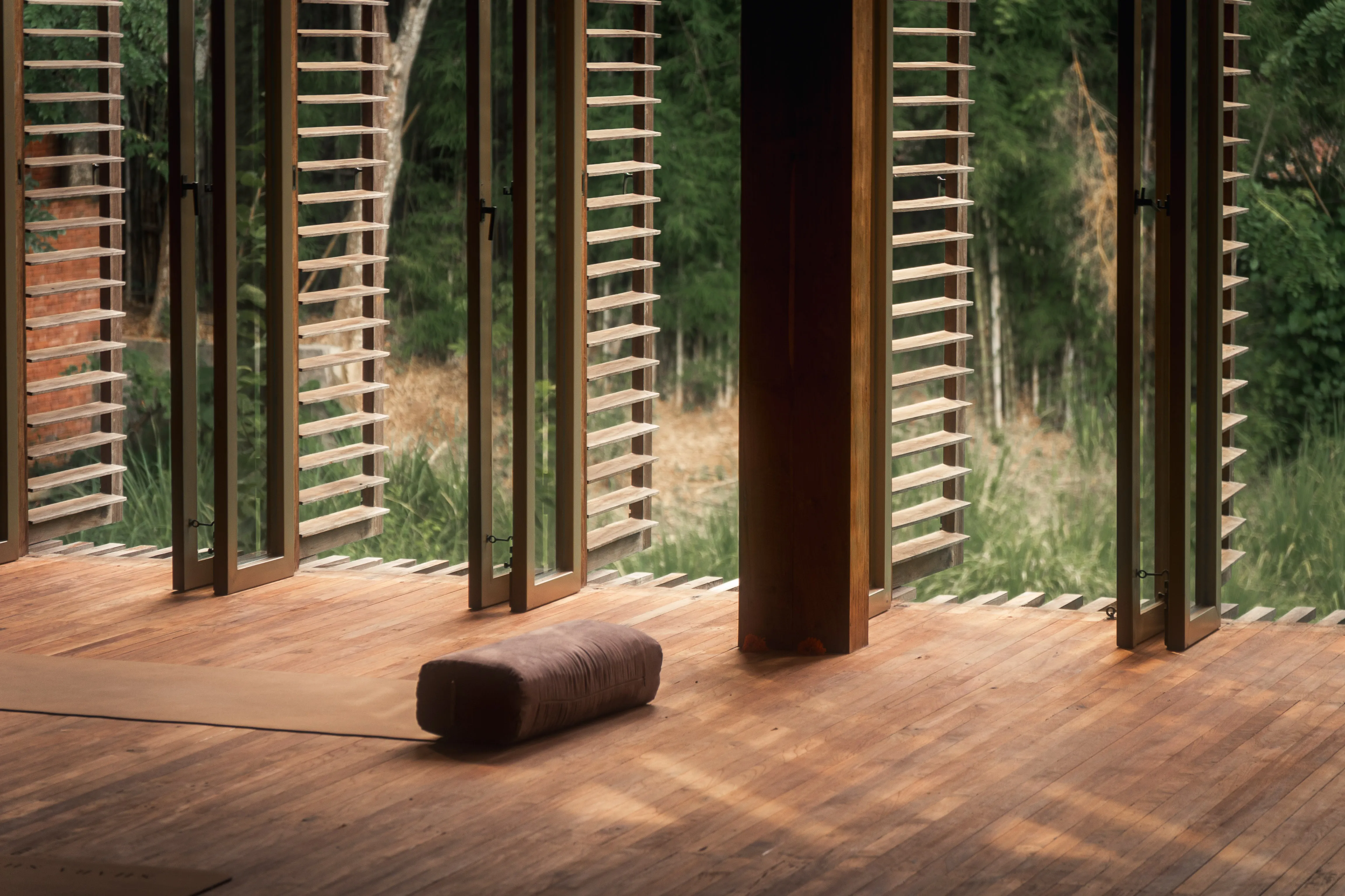 Empty yoga mat and bolster set on polished teak wood flooring inside an open-air studio with louvered doors at The Shaba Shala.