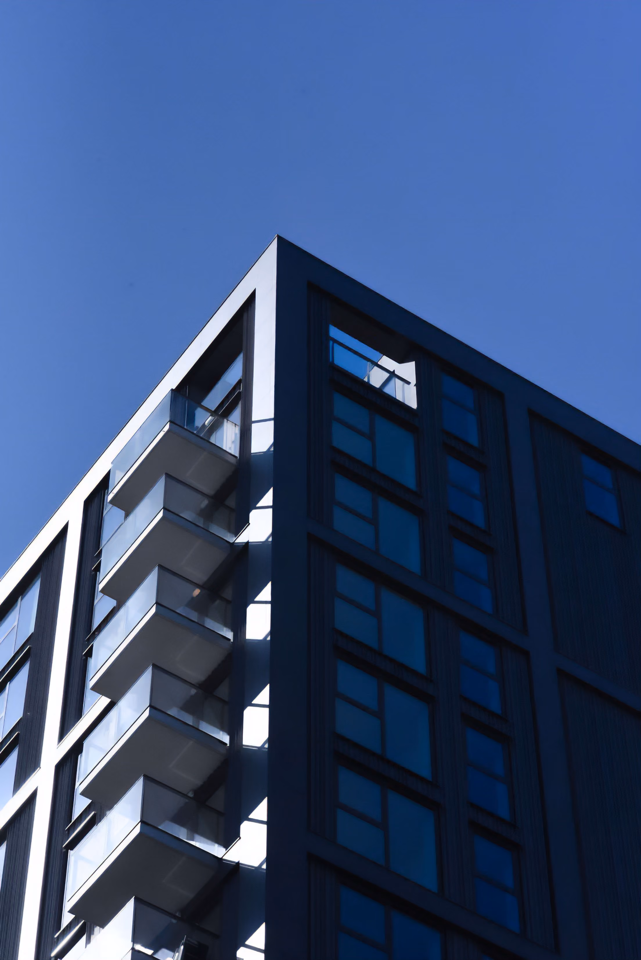 Façade d'un immeuble moderne avec balcons en verre et grandes fenêtres sous un ciel bleu clair.