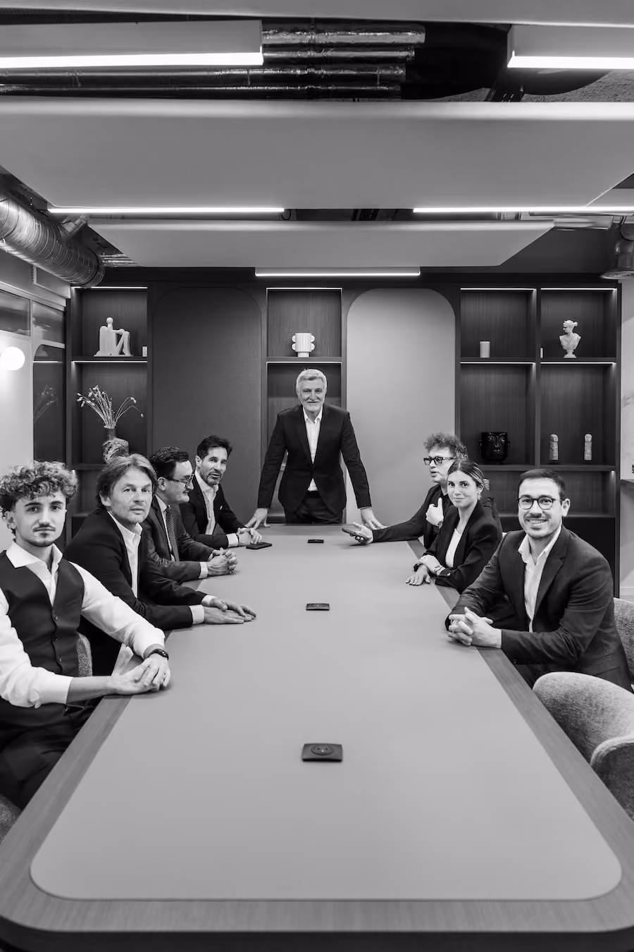 Sept professionnels en costume assis et debout autour d'une grande table de réunion dans un bureau moderne.